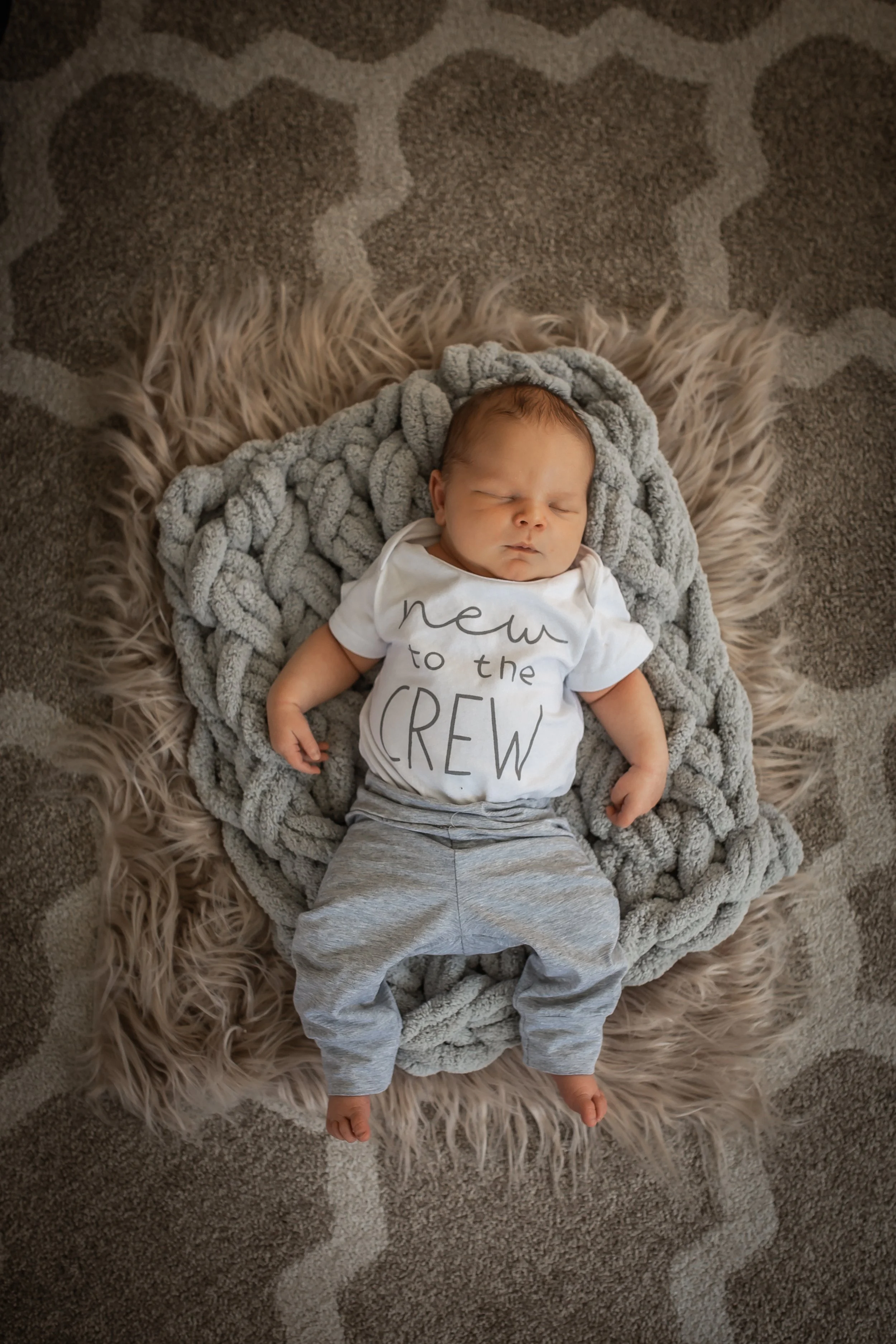 Newborn baby sleeping on a textured blanket, wearing a "new to the crew" onesie and gray pants, on a patterned rug.