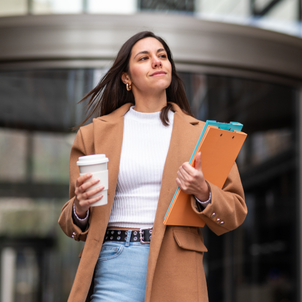 Woman walking outdoors with a coffee and folders, looking confident and relaxed.