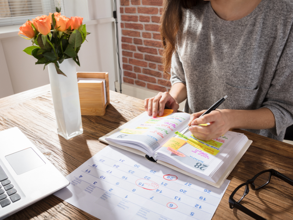 Woman organising her weekly schedule in a planner, with a laptop and flowers on the table.