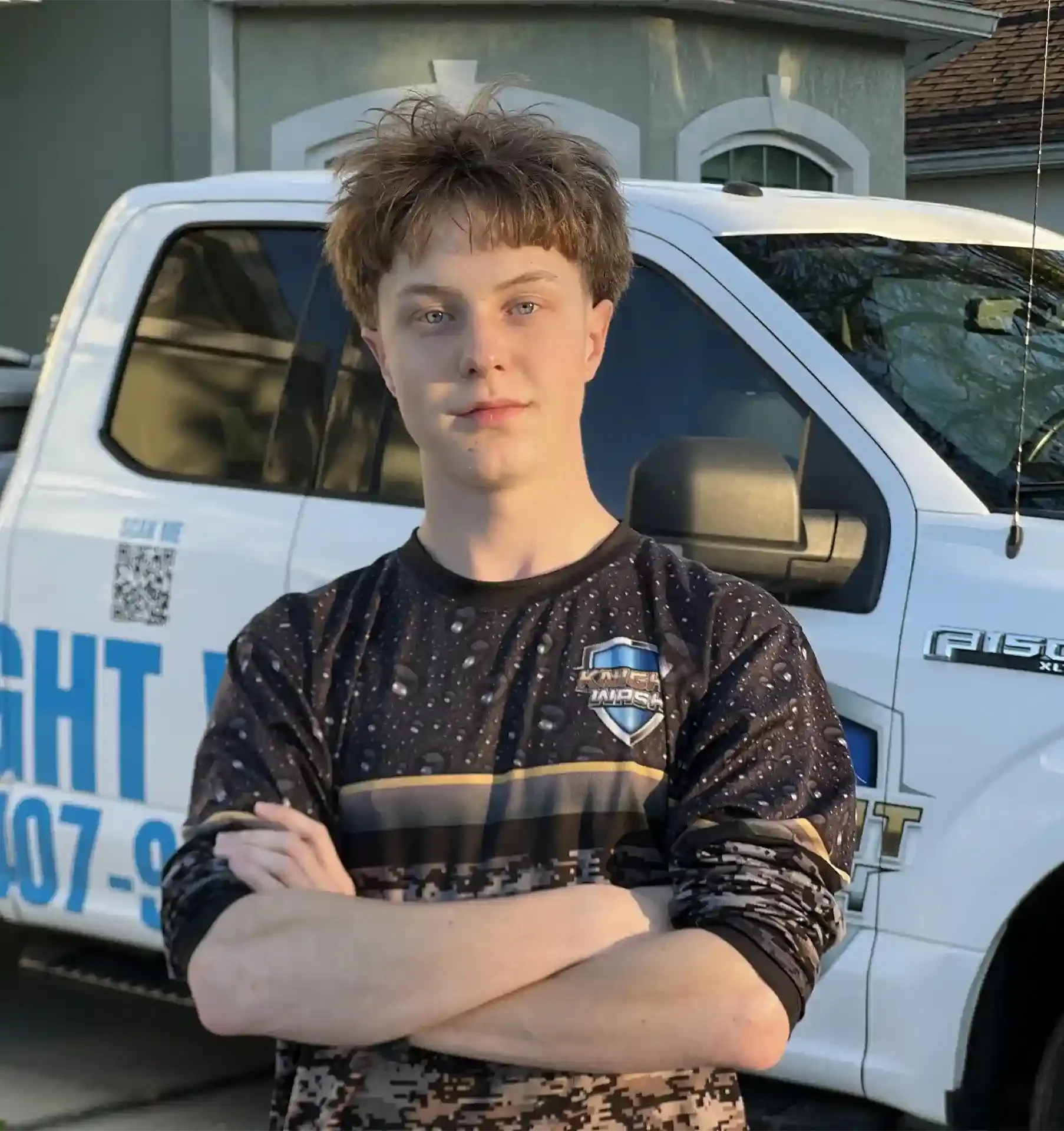 A knight wash technician with short, messy hair, standing with arms crossed in front of a white pickup truck with a company logo on the side, in an outdoor residential setting.