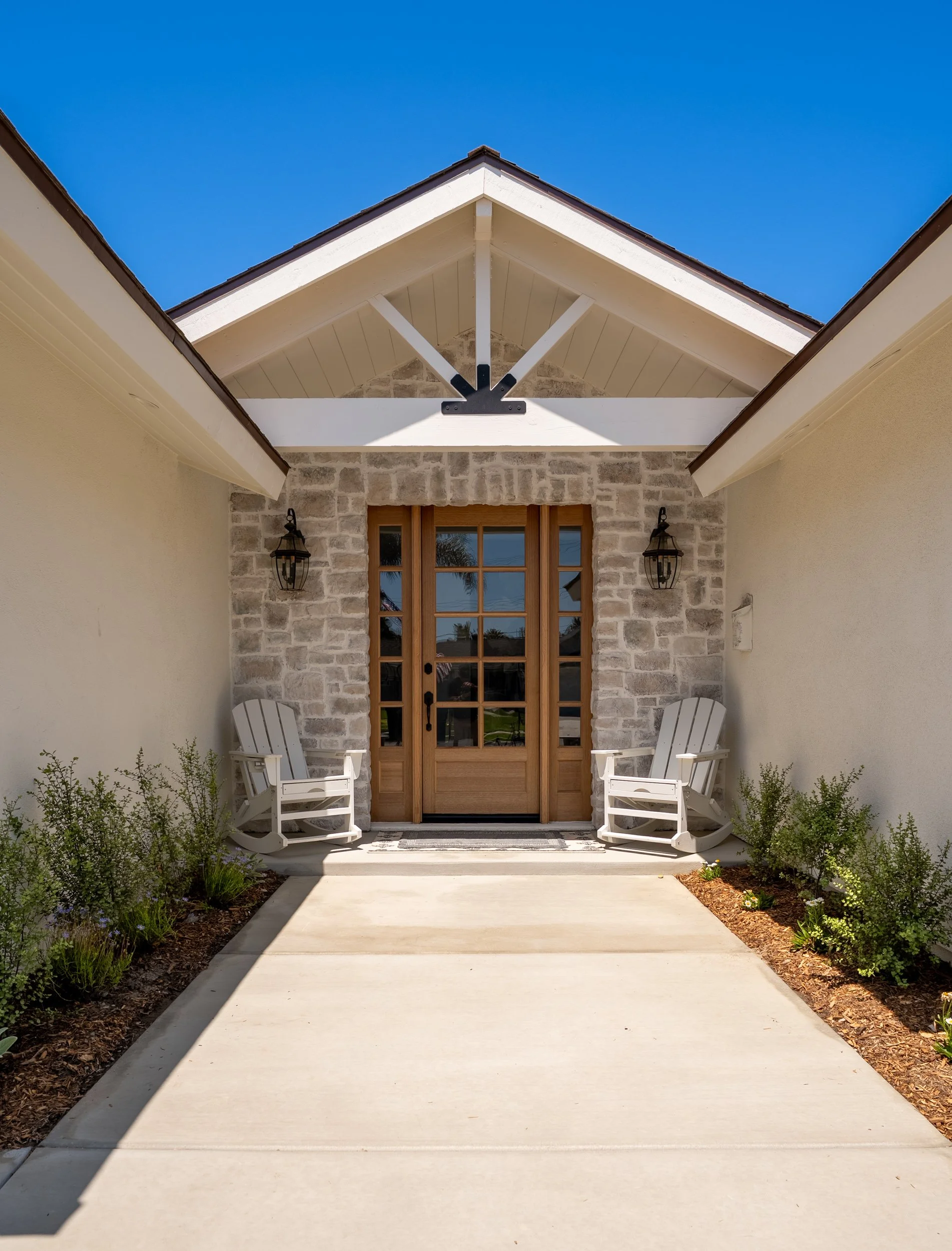 Front porch view with a concrete walkway leading to a wooden door, flanked by two white rocking chairs, with stone and stucco walls, and a gable roof with white trim and black brackets.