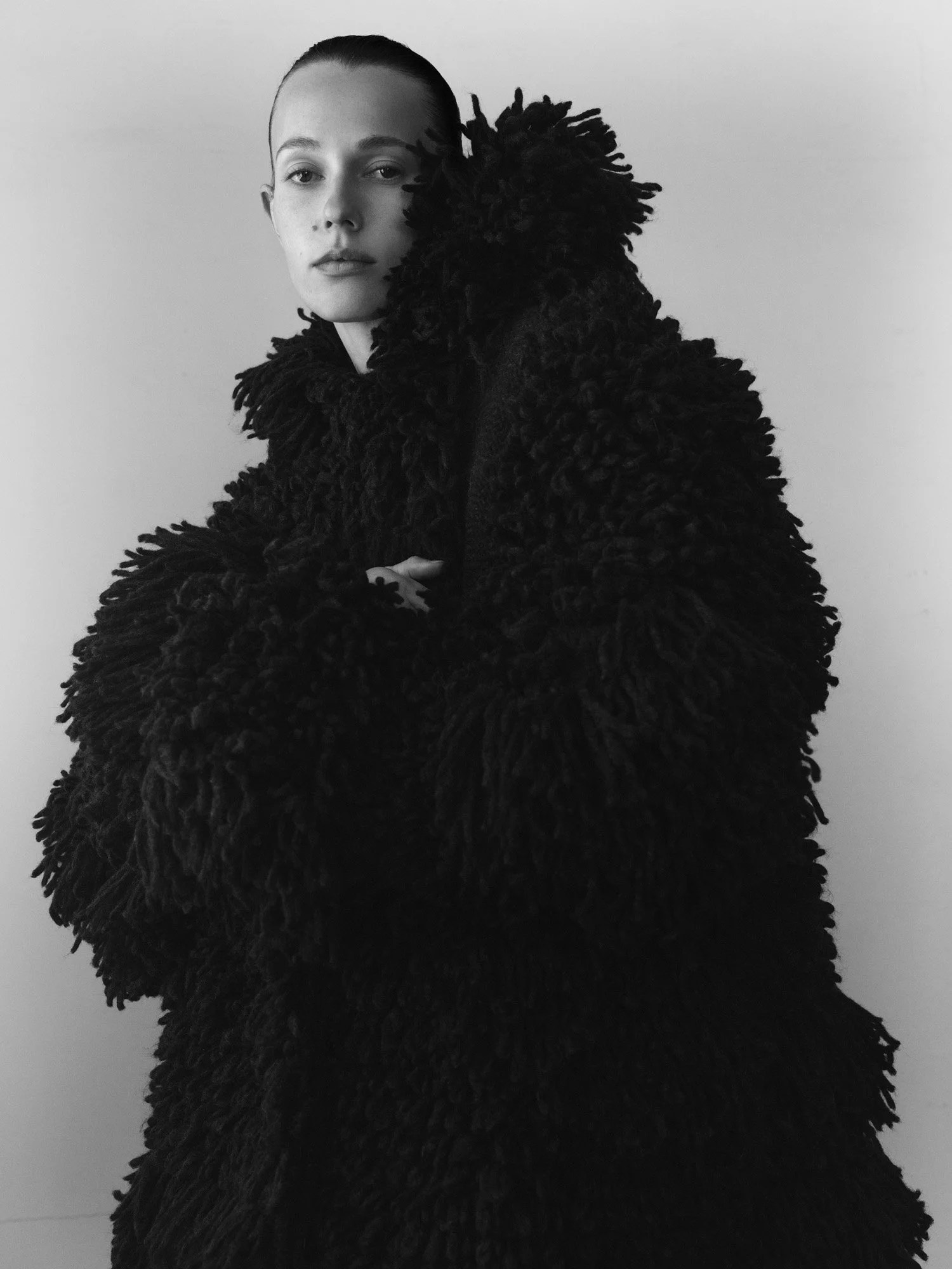 Black and white photograph of a young woman with a shaved head wearing a large, fluffy, textured black coat, standing against a plain background.
