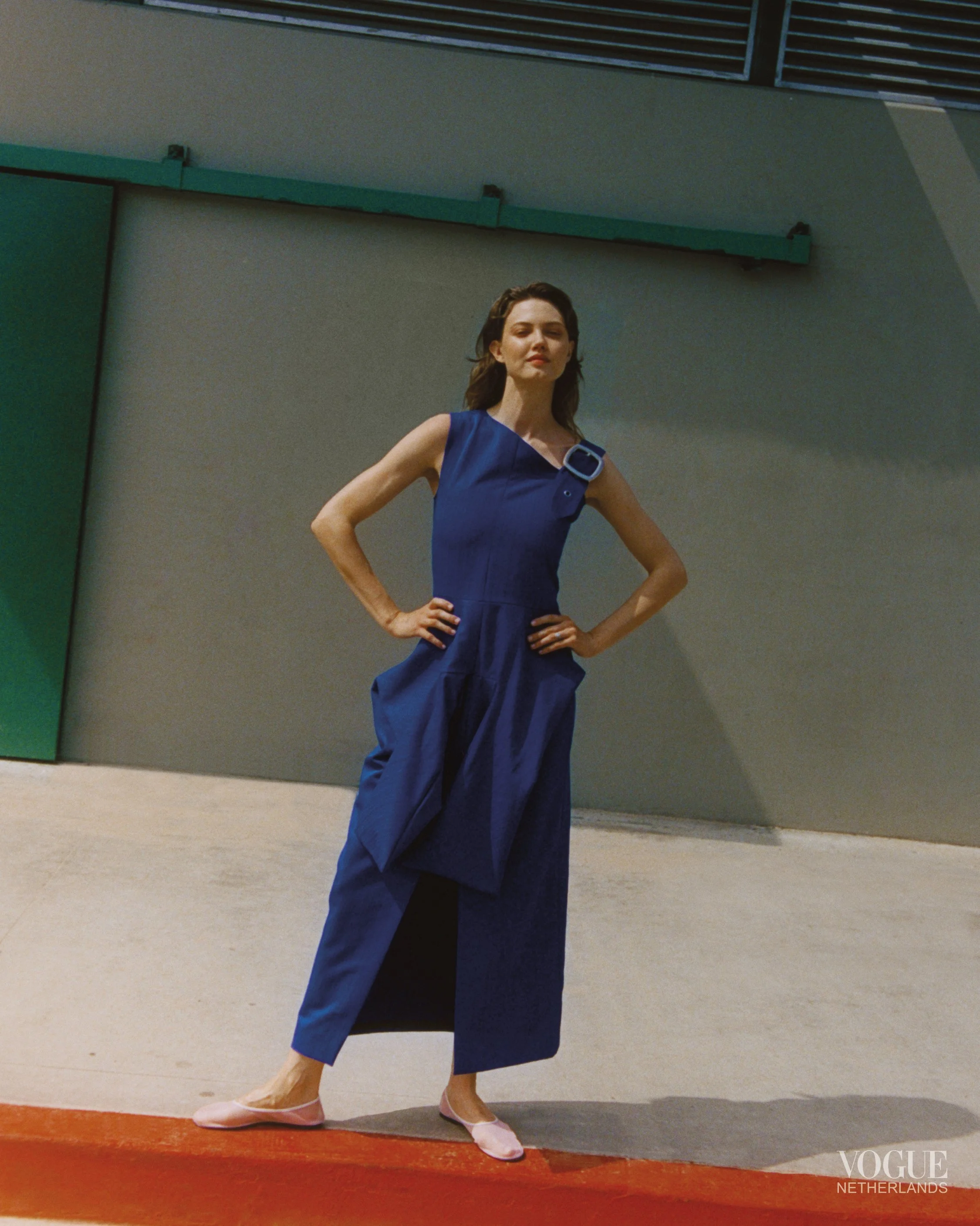 A woman wearing a blue dress standing confidently with her hands on her hips on a runway, with a plain background and the Vogue Netherlands logo in the bottom right corner.