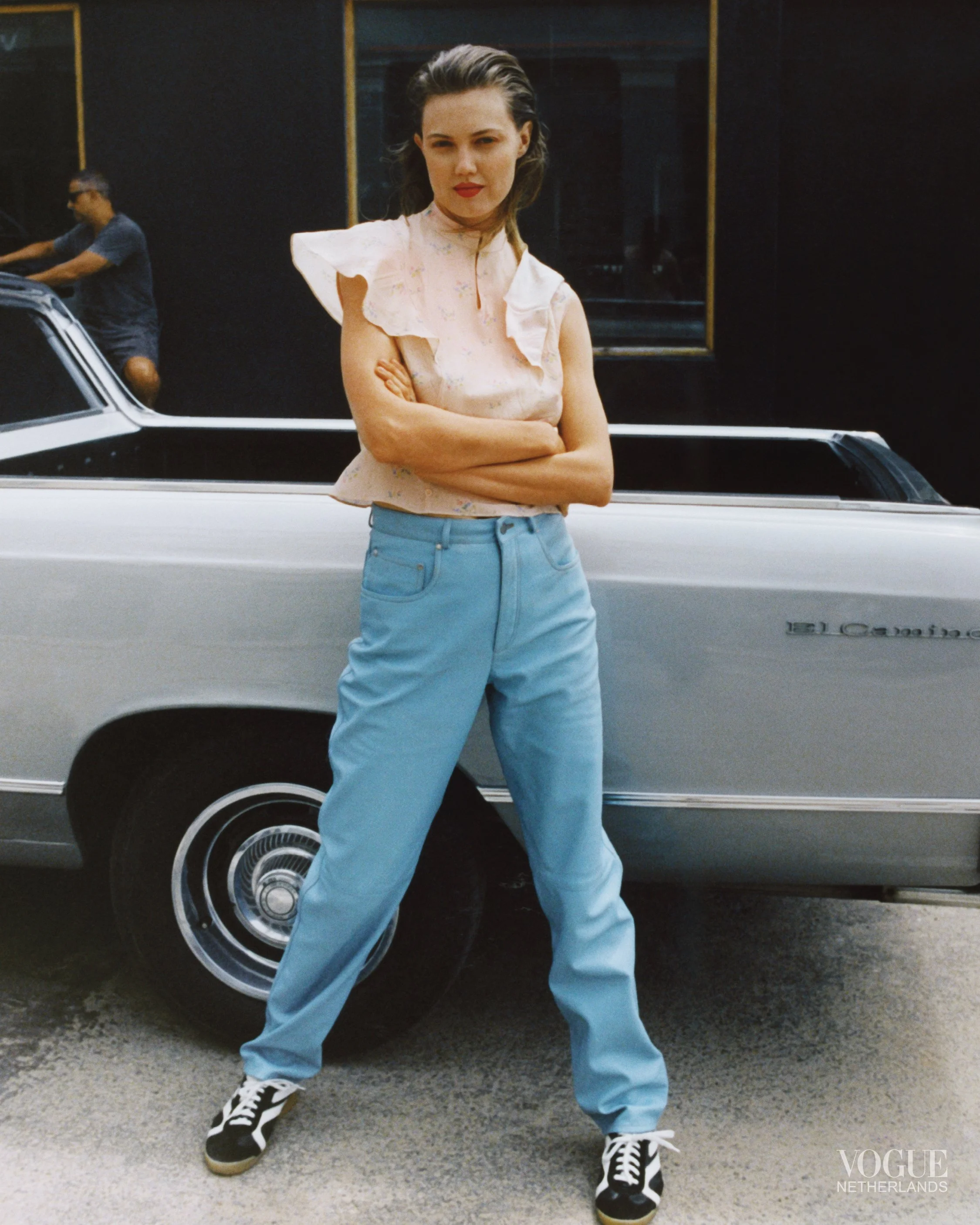 A woman with dark hair, wearing a light pink ruffled blouse, light blue jeans, and sneakers, stands with arms crossed in front of a vintage silver car. A person in the background is riding a bicycle.