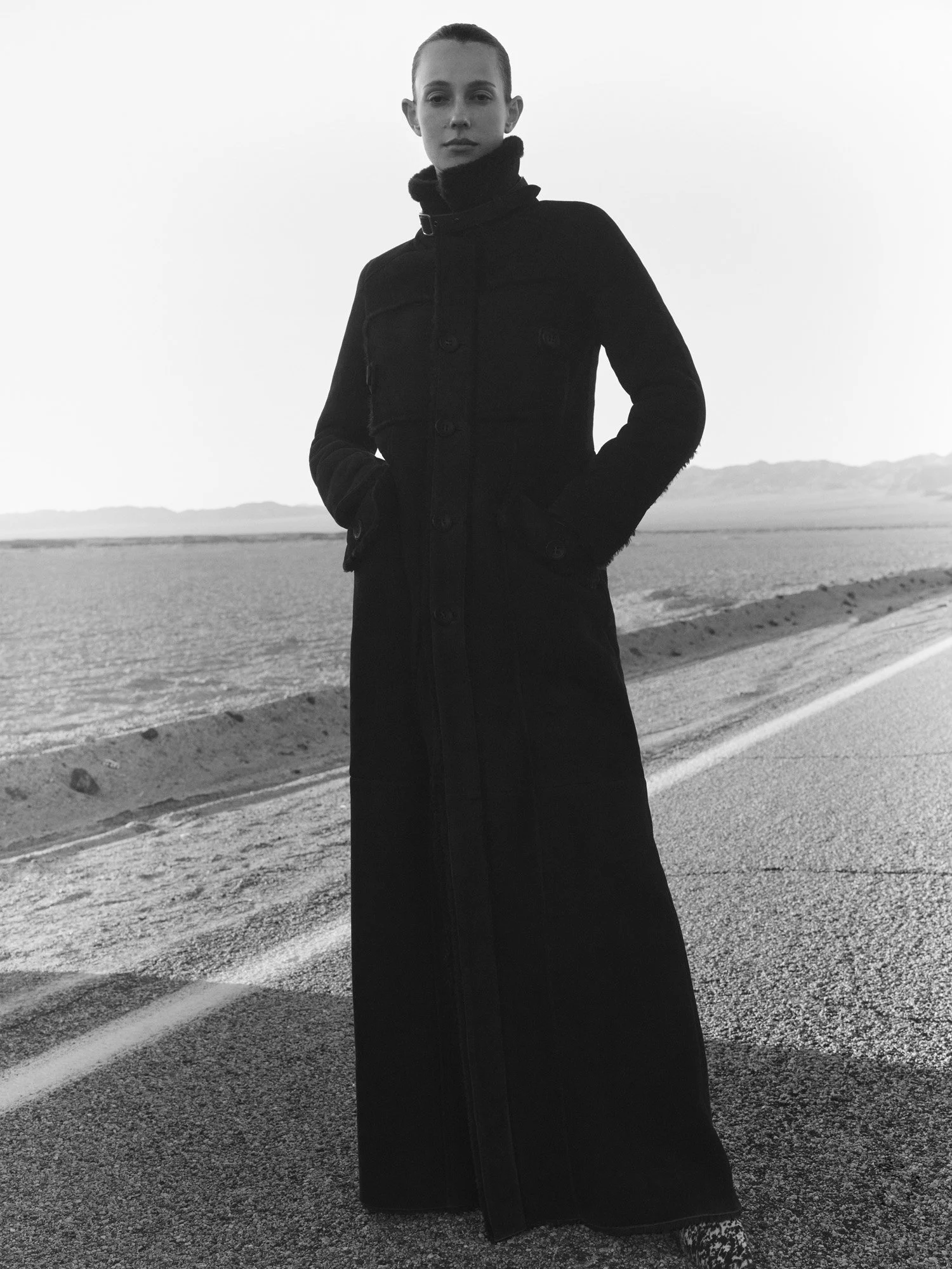 A woman stands on a road in a desert landscape wearing a long black coat with a high collar, hands in pockets, looking at the camera in black and white.
