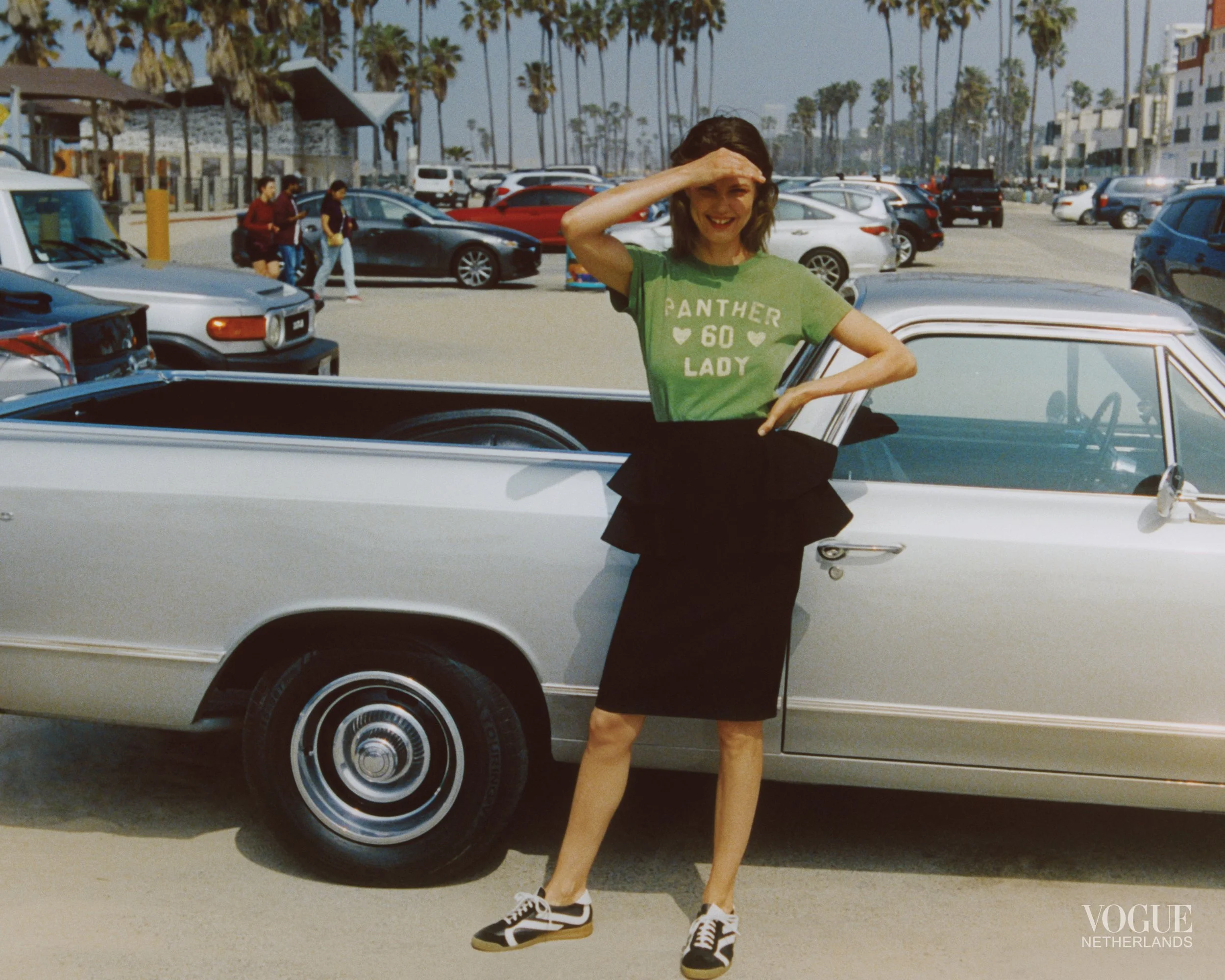 A woman standing next to a classic silver car at a parking lot, saluting with her right hand and smiling.