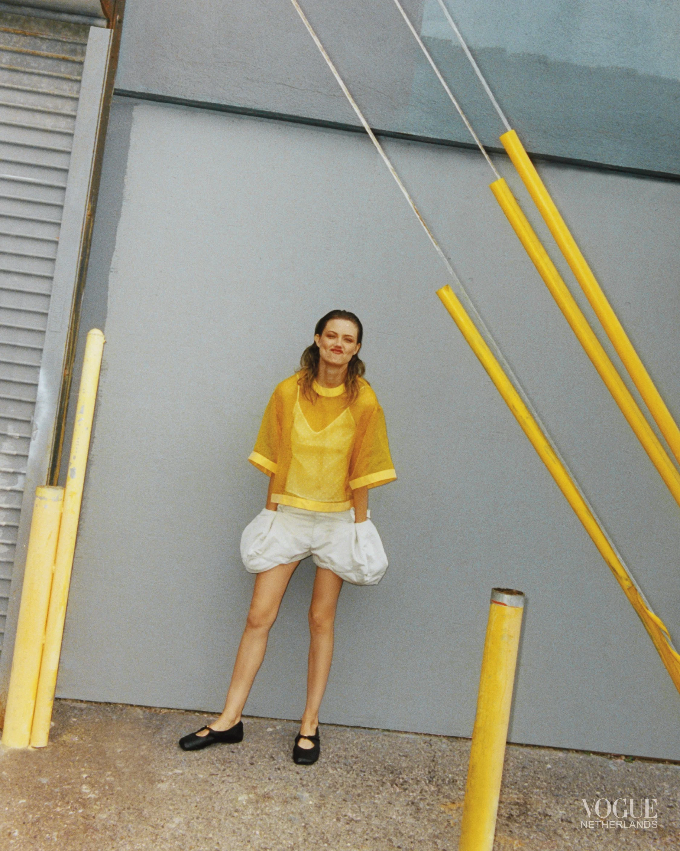 A woman standing outdoors against a plain gray wall, wearing a yellow sheer top, white puffed shorts, and black shoes, with yellow poles nearby, and a small 'Vogue Netherlands' logo in the bottom right corner.