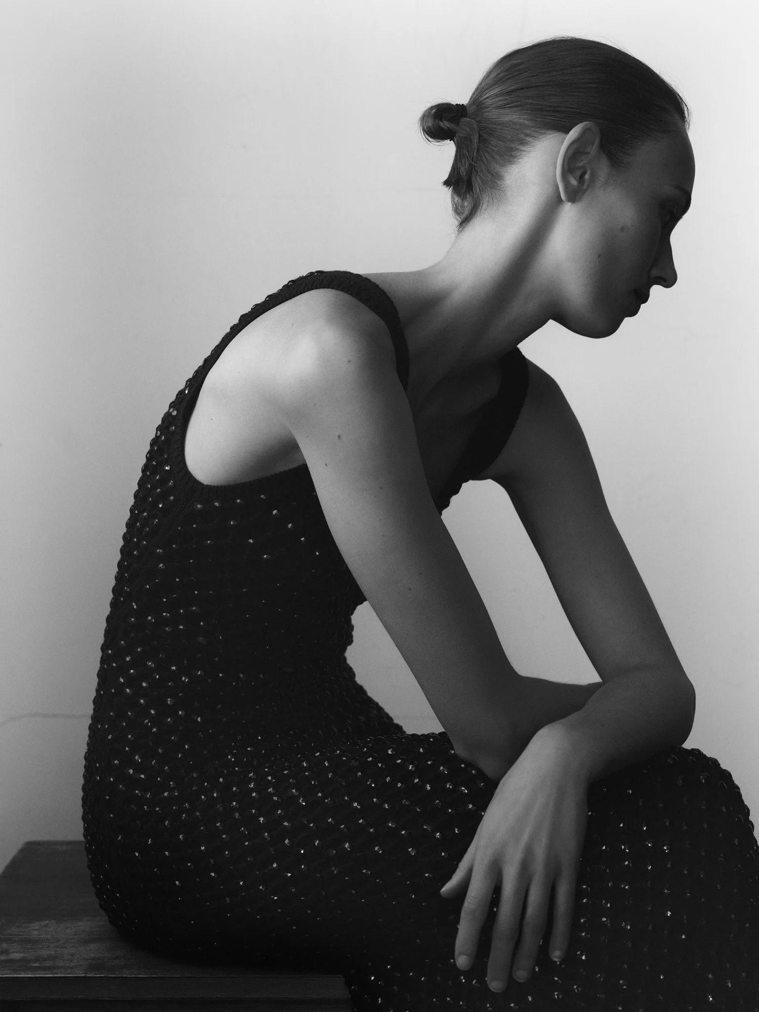 Black and white photo of a woman sitting on a bench with her head turned downward, wearing a sleeveless dress with small shiny dots and her hair tied back.