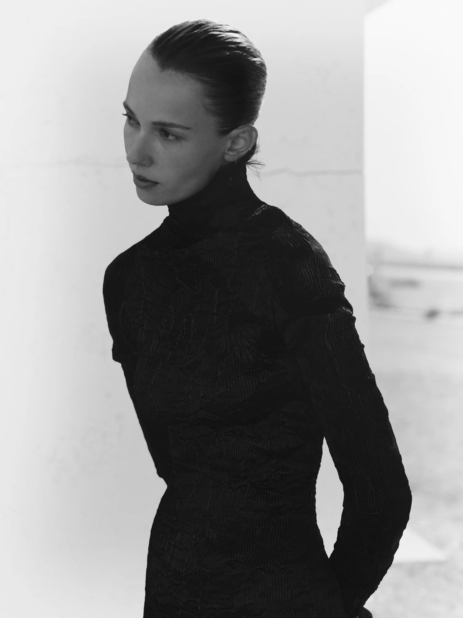 Black and white photo of a woman with slicked-back hair wearing a long black textured dress, standing indoors with a plain background.