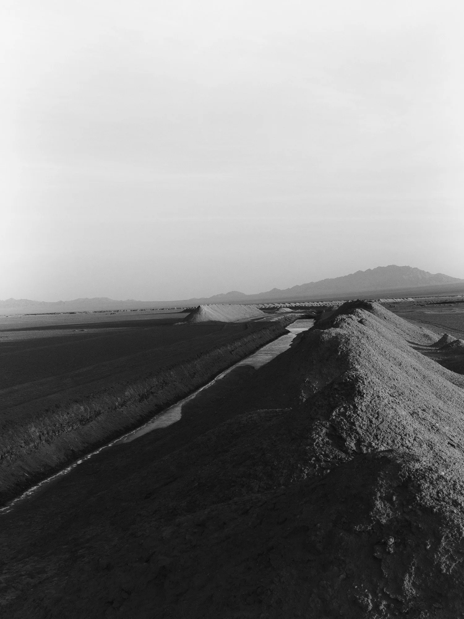 Black and white landscape of a desert with mounds of salt and a narrow canal running through the salt flats, mountains in the background.