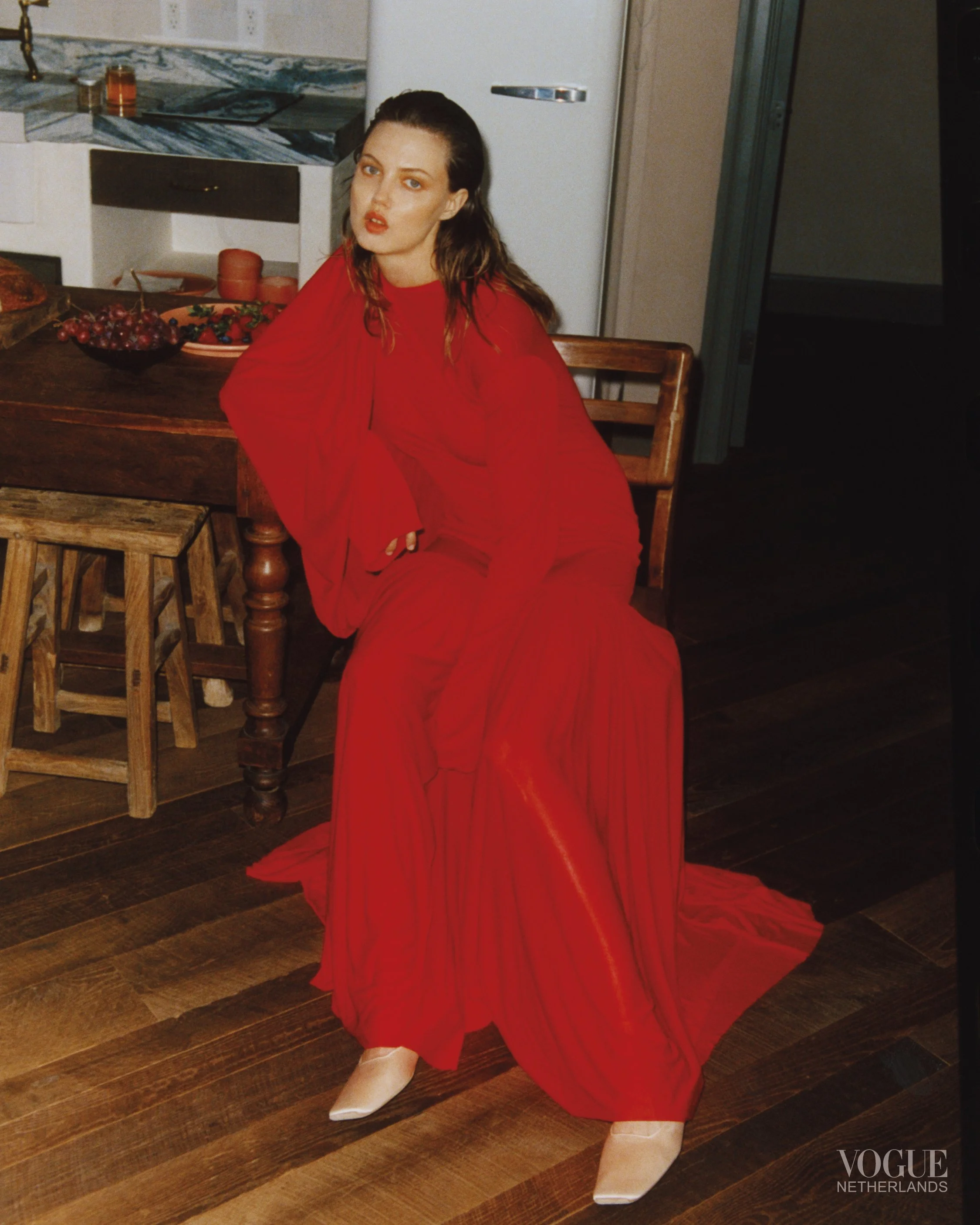 A woman dressed in a long, flowing red gown with pleats, sitting on a wooden chair at a kitchen table with bowls of fruit, in a rustic kitchen setting. She is wearing beige pointed shoes and has dark, wavy hair.