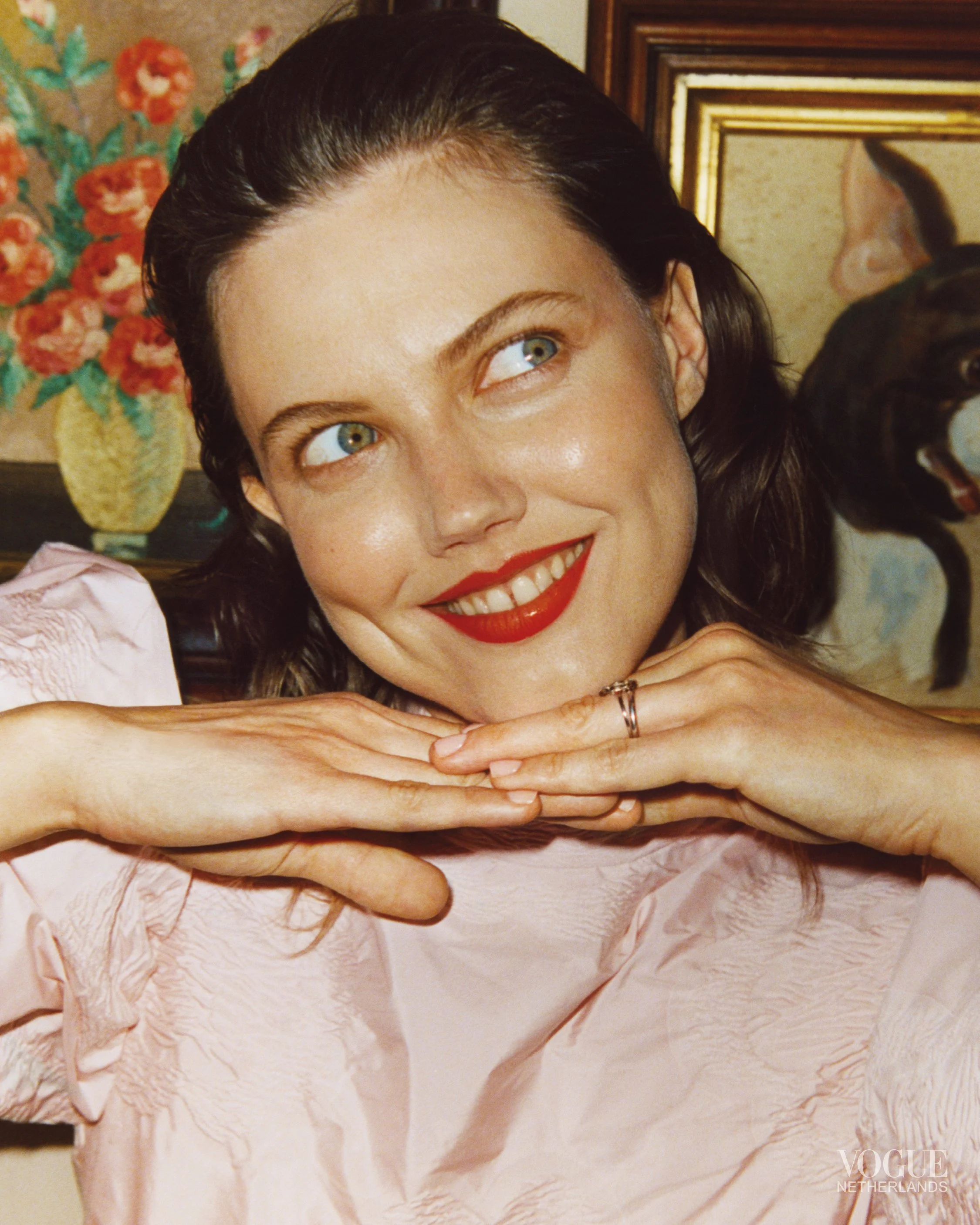 Close-up of a woman with blue eyes and dark hair smiling, wearing red lipstick, resting her chin on her folded hands, with a pink blouse. Background includes a framed picture, a vase with orange flowers, and a dog with its mouth open.