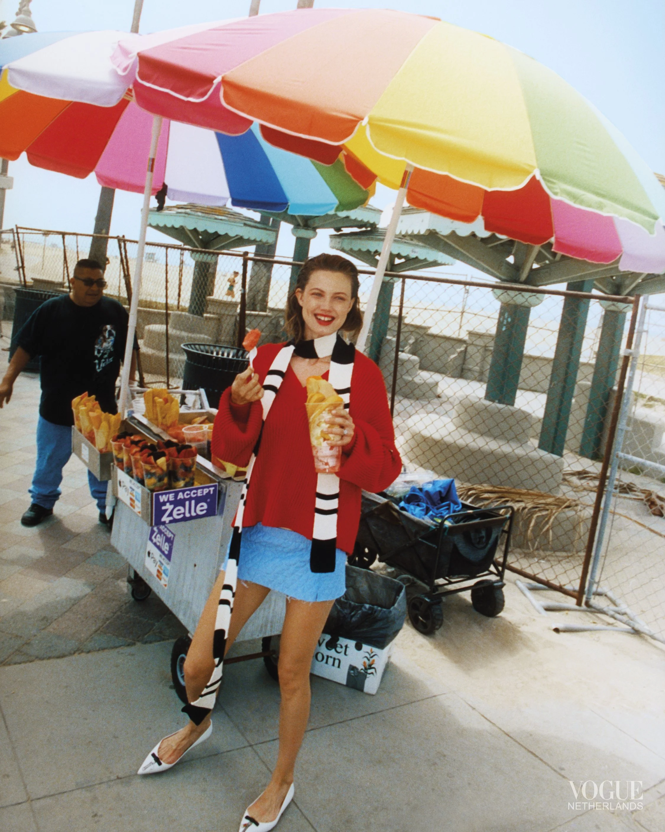 A woman in a red sweater, blue skirt, and white heels holding a snack at a hot dog stand with a colorful umbrella. Behind her, a man is near the stand, and there are various supplies and signs around the stand.