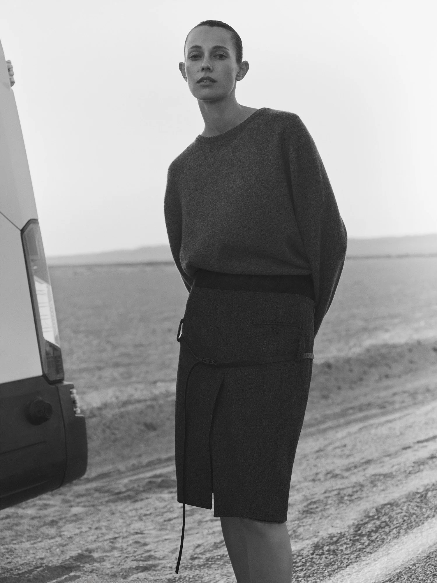 A woman in a dark sweater and skirt standing outdoors on a dirt road, with a vehicle partially visible on the left side of the image, and a barren landscape background.