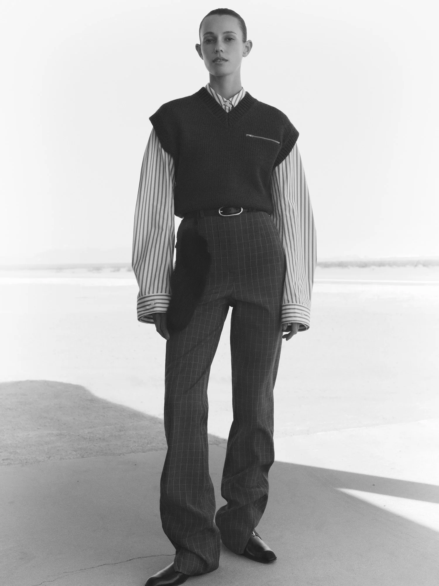 Black and white photo of a woman standing outdoors on a beach, wearing a sleeveless sweater vest over a striped shirt, checkered pants, and dark shoes.
