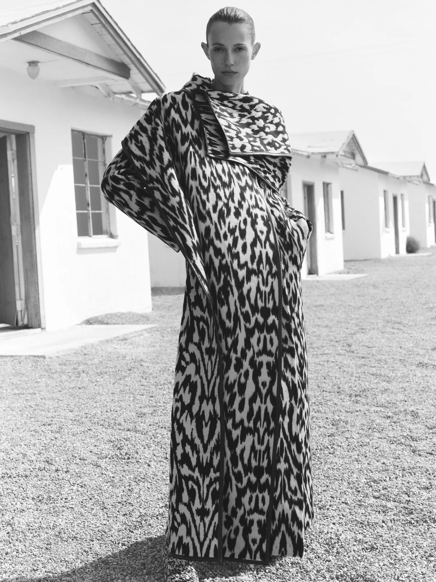 A woman standing outdoors wearing a long, animal print coat with a large collar, in front of a row of small houses, black-and-white photo.