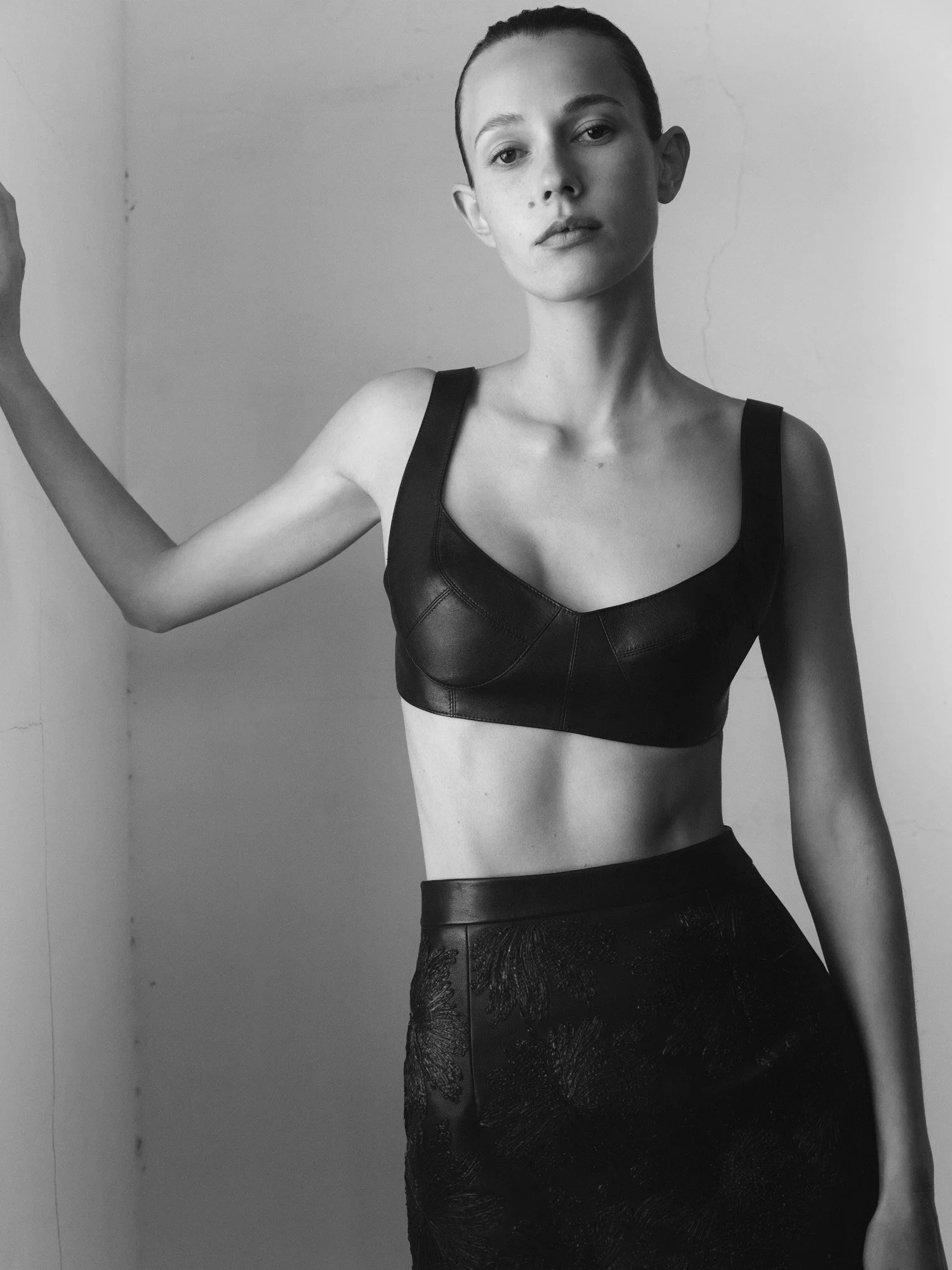 Black and white photograph of a young woman with slicked-back hair, wearing a black leather crop top and high-waisted skirt, standing against a plain wall.