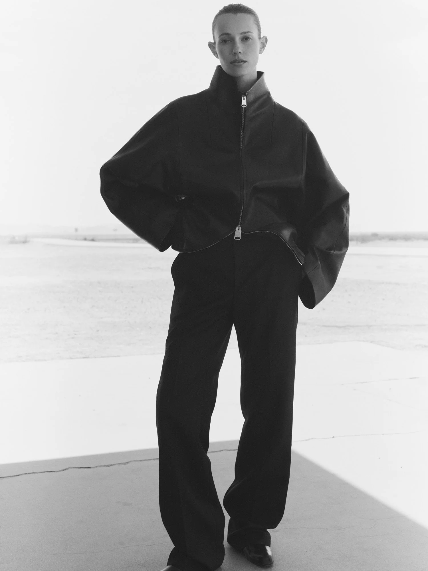 A woman with slicked-back hair wearing a black leather jacket, loose black pants, and black shoes, standing outdoors with a plain background.