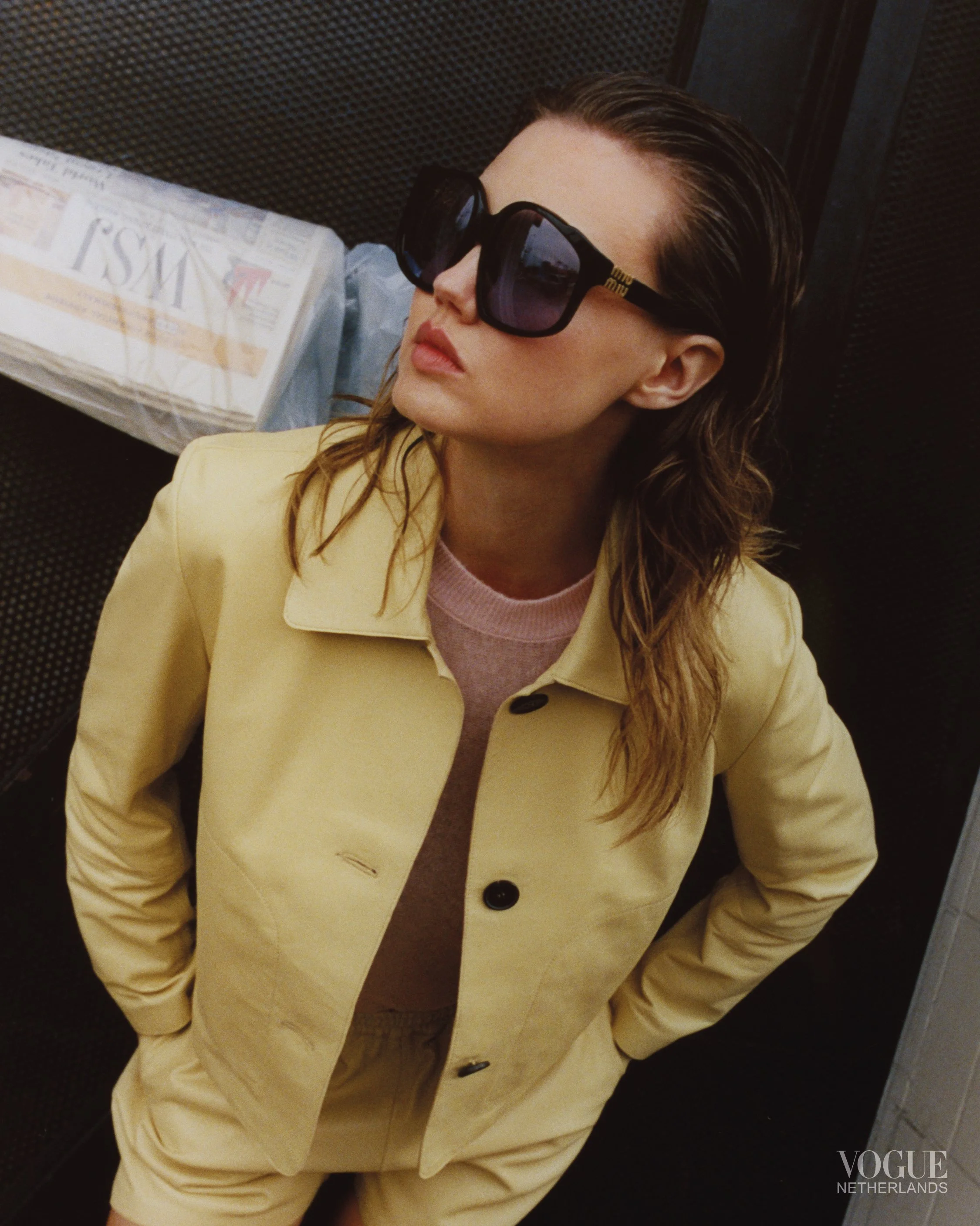 A woman with brown wavy hair wearing large black sunglasses, a beige coat, and a pink shirt, standing with her hands on her hips in an indoor location.