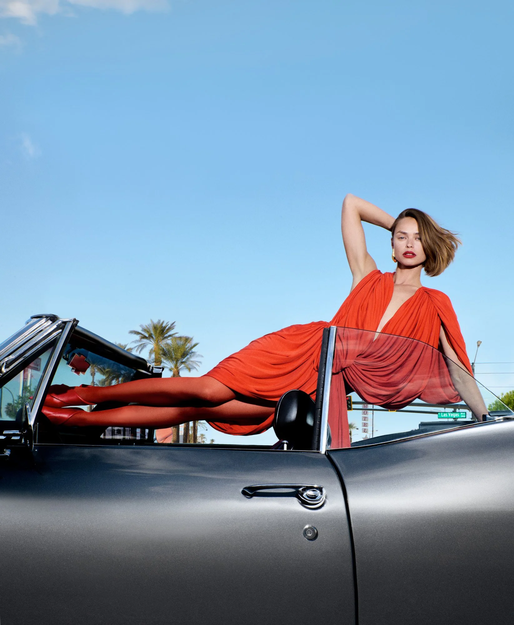 A woman in an orange dress lying on the side of a black convertible car parked outdoors with palm trees and a clear blue sky in the background.