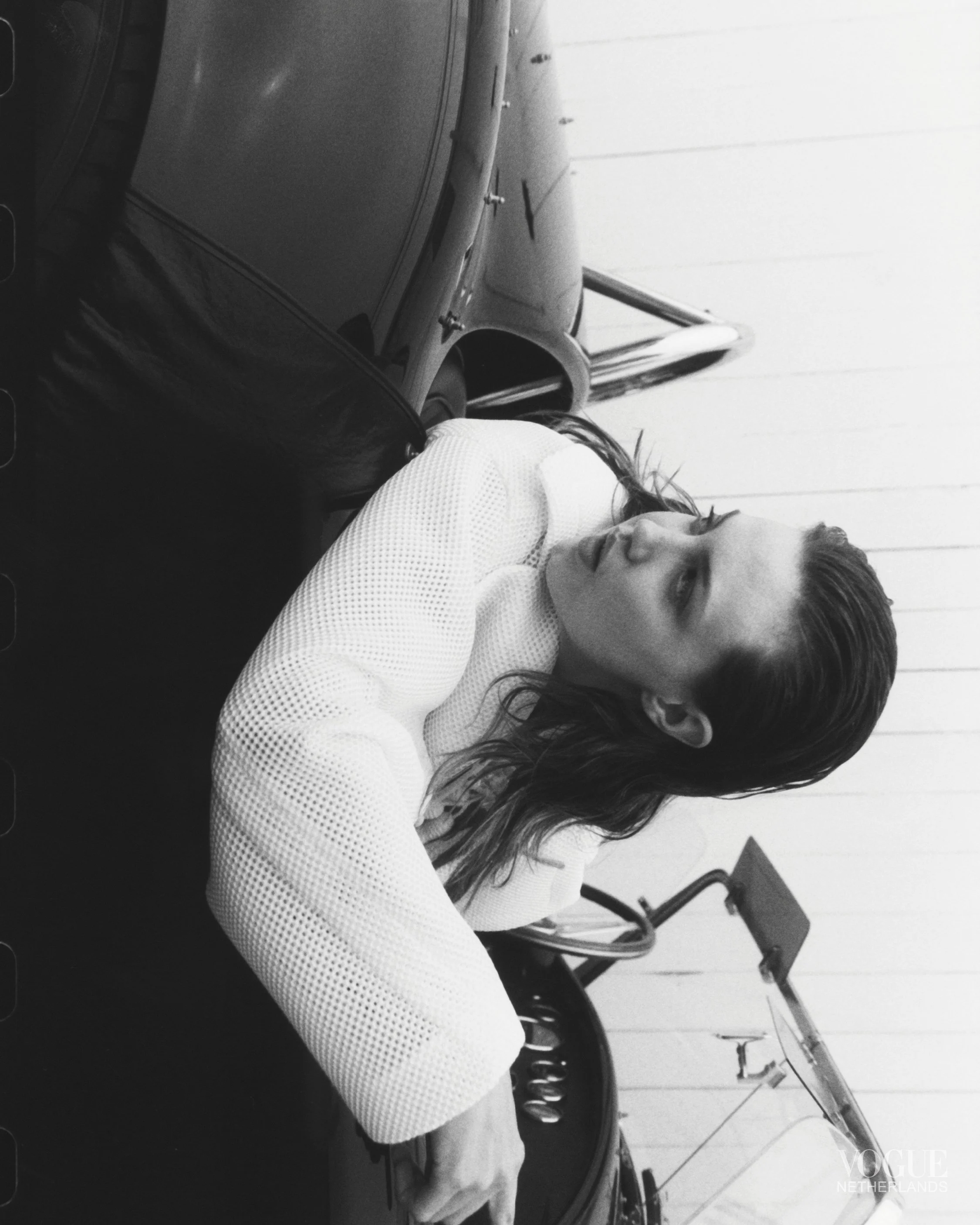 A woman with wet hair wearing a white textured top sitting with her arms crossed on a table, looking sideways in a black-and-white photo with a boat and a wooden wall in the background.