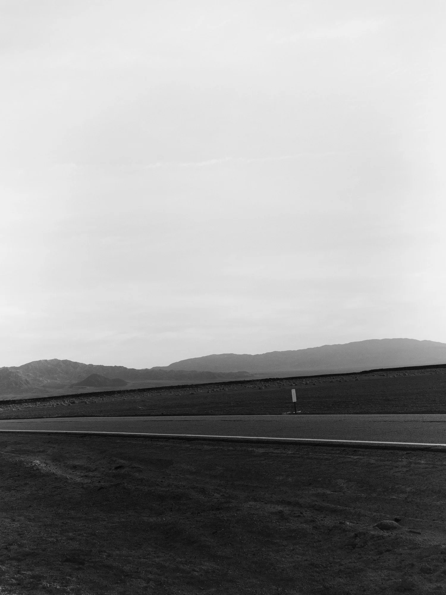 Black and white landscape with a road in the foreground, distant mountains, and a cloudy sky.