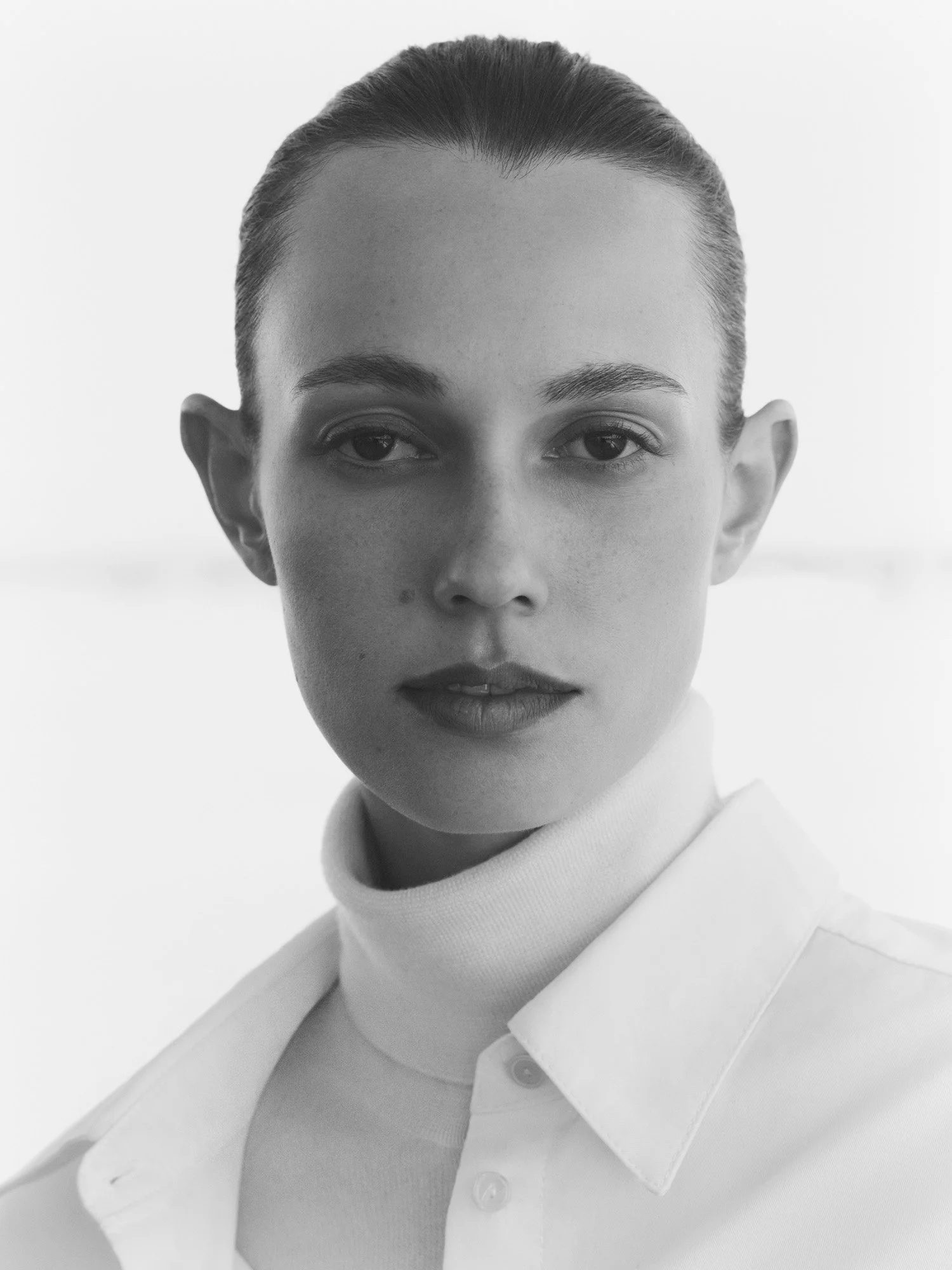 Black and white portrait of a woman with her hair pulled back, wearing a turtleneck and a button-up shirt, looking directly at the camera.