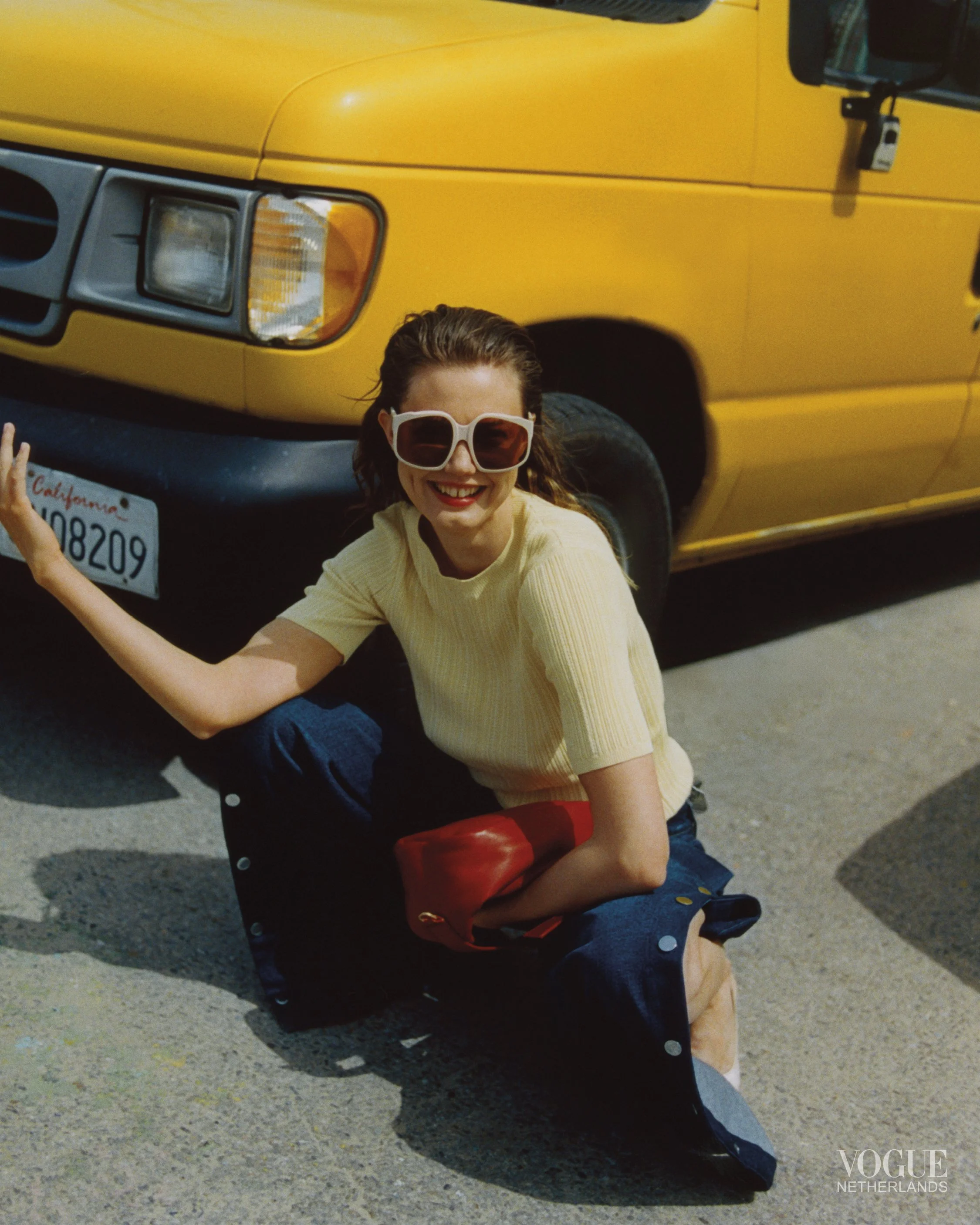 A woman with sunglasses and a yellow shirt squatting on the ground next to a yellow vehicle, smiling.