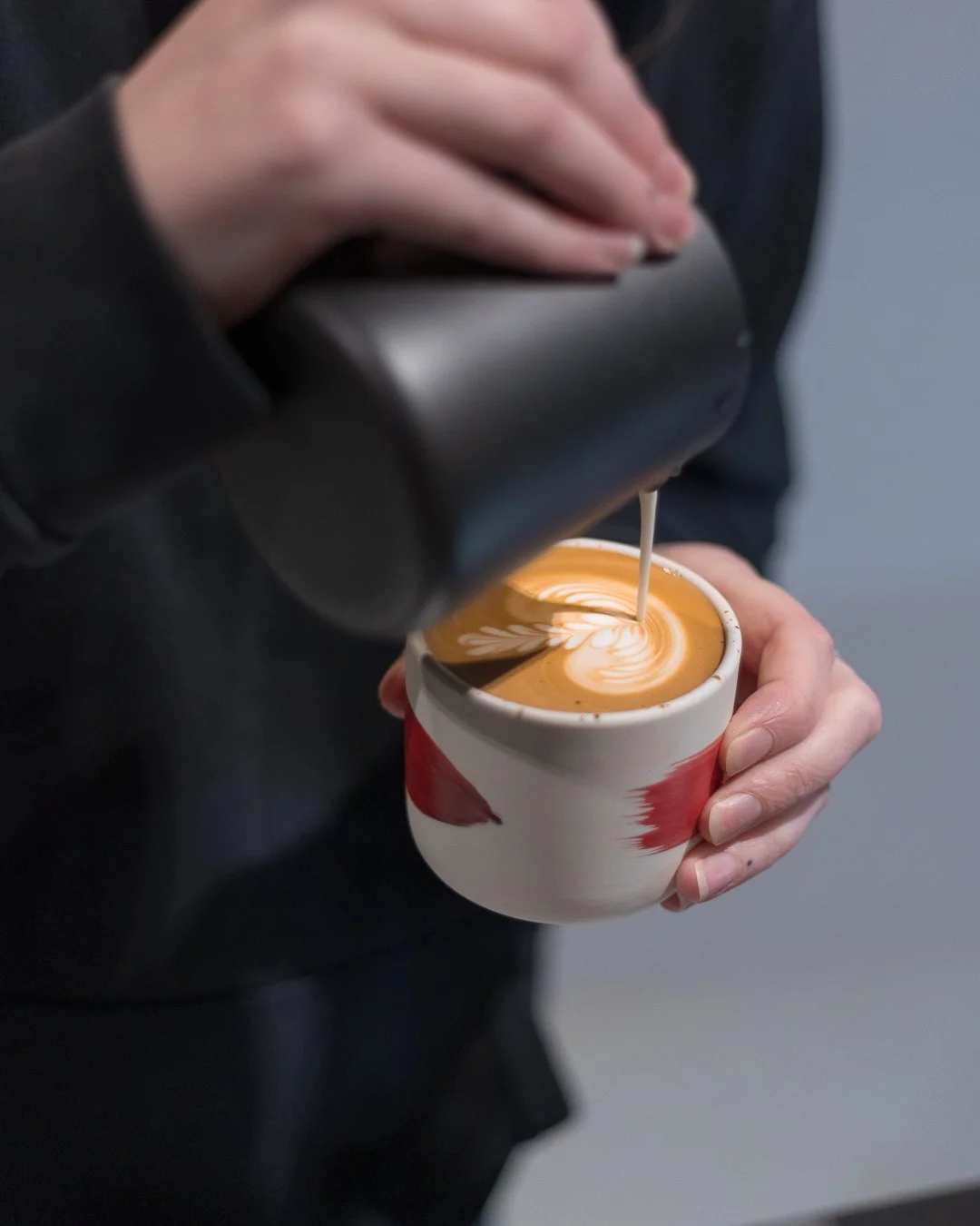 Person pouring steamed milk into a latte with latte art in a ceramic mug.
