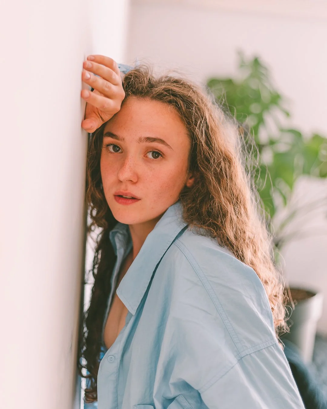 A young woman with curly hair and freckles wearing a light blue shirt, leaning against a white wall, looking at the camera with a neutral expression.