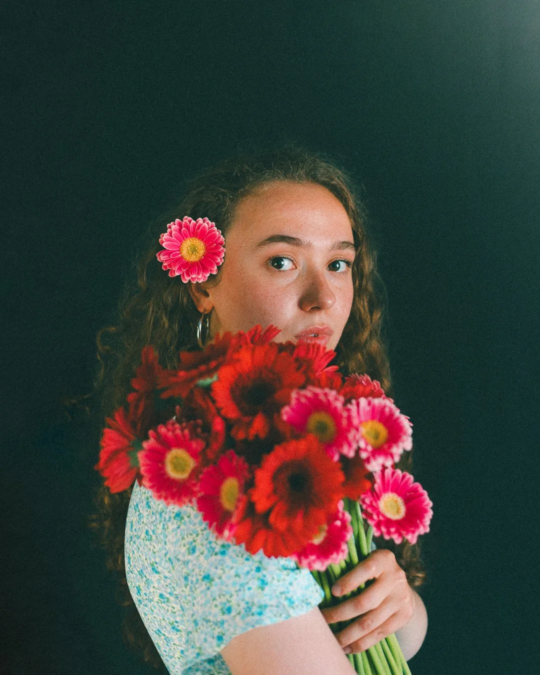 A young woman with curled hair and hoop earrings holds a bouquet of pink and red flowers, with a pink flower tucked behind her ear, against a dark background.