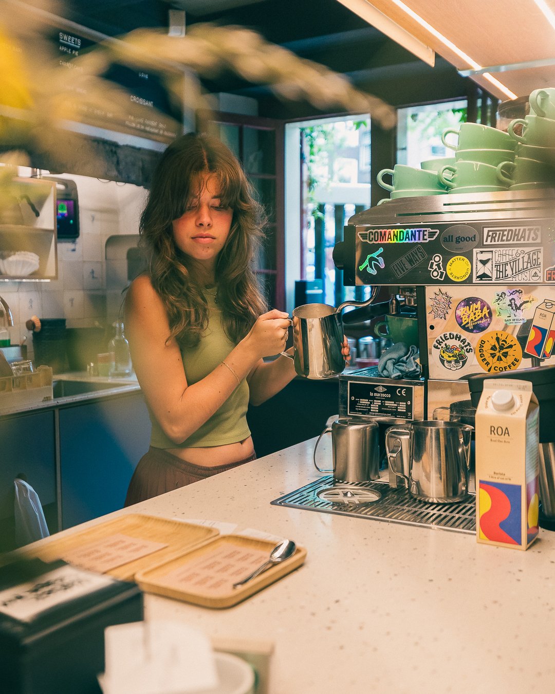 A young woman with long, wavy hair standing at a coffee shop counter pouring hot water from a metallic kettle into a cup. The counter has a cream-colored surface with a menu, a spoon, and various coffee-making equipment. Behind her, there are colorfu