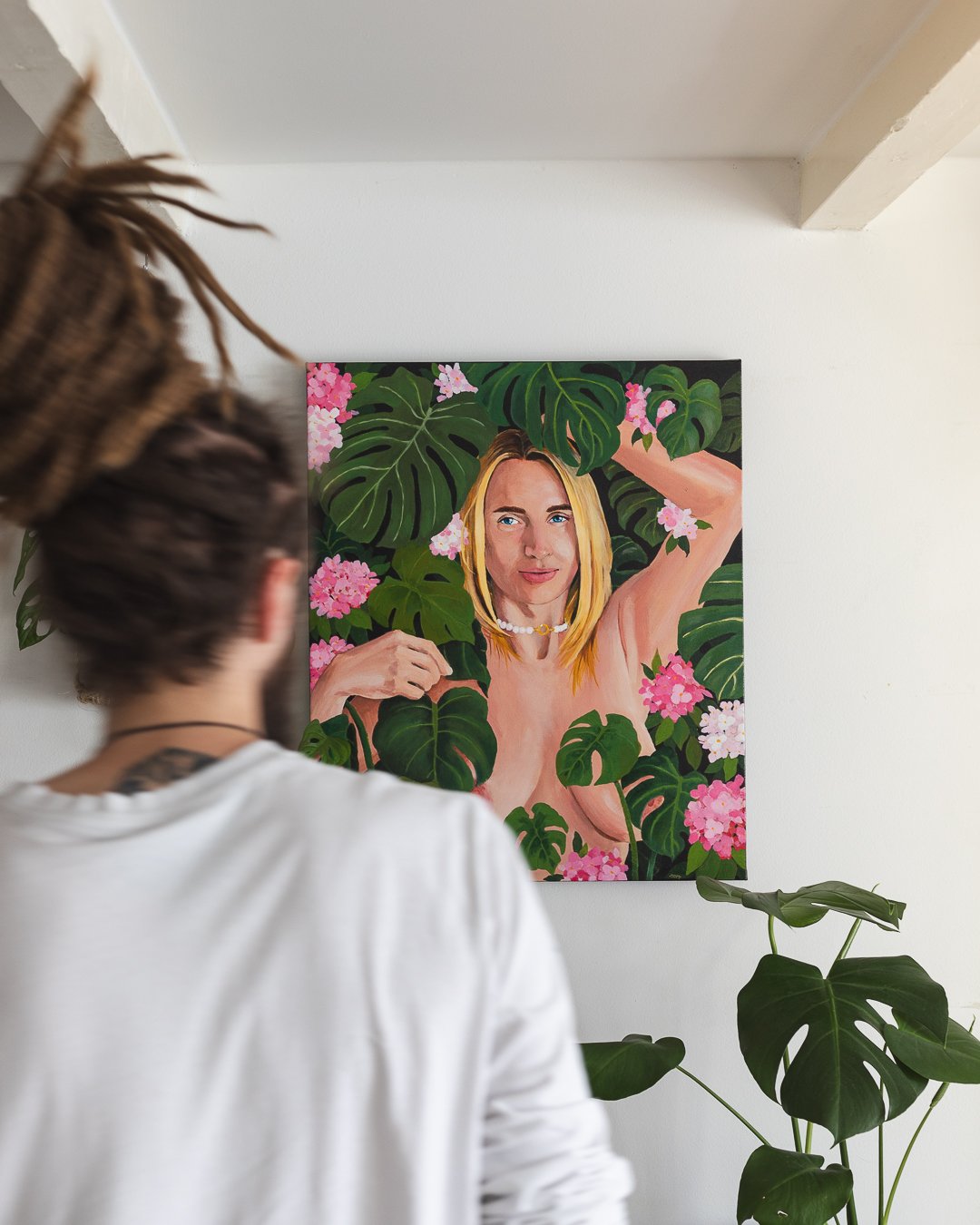 Person with dreadlocks and a choker necklace observing a colorful portrait of a blonde woman with blue eyes surrounded by lush green leaves and pink flowers on a gallery wall.