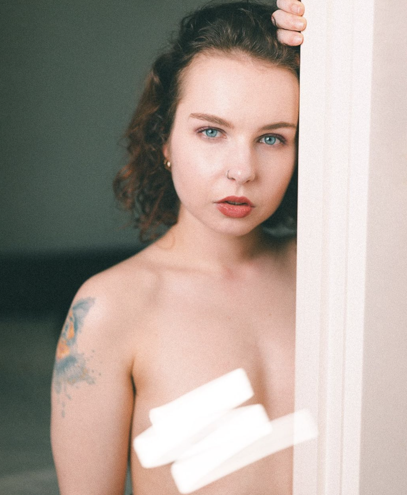 A woman with short curly hair and blue eyes looking at the camera, partially hidden behind a door or wall, with a tattoo on her shoulder and holding two rolled-up white towels.