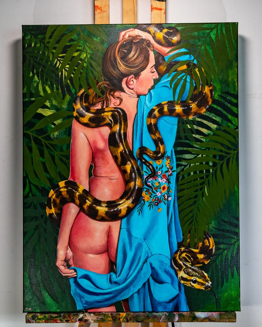 An artwork depicting a woman with brown hair hiding her face with her eyes closed, joined by a large snake with yellow and black markings wrapped around her body, set against a background of green tropical leaves.