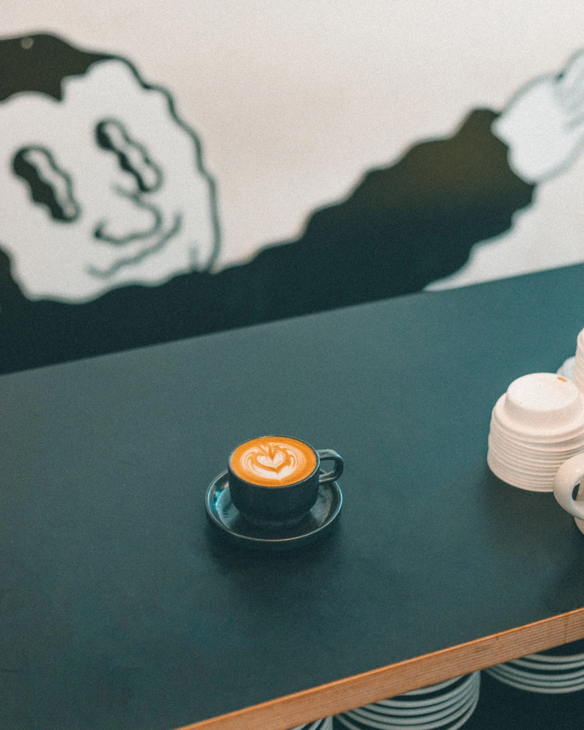 Close-up of a cup of coffee with latte art on the foam, placed on a silver saucer on a dark green table. In the background, there are white paper cups stacked together, and part of a table edge is visible.