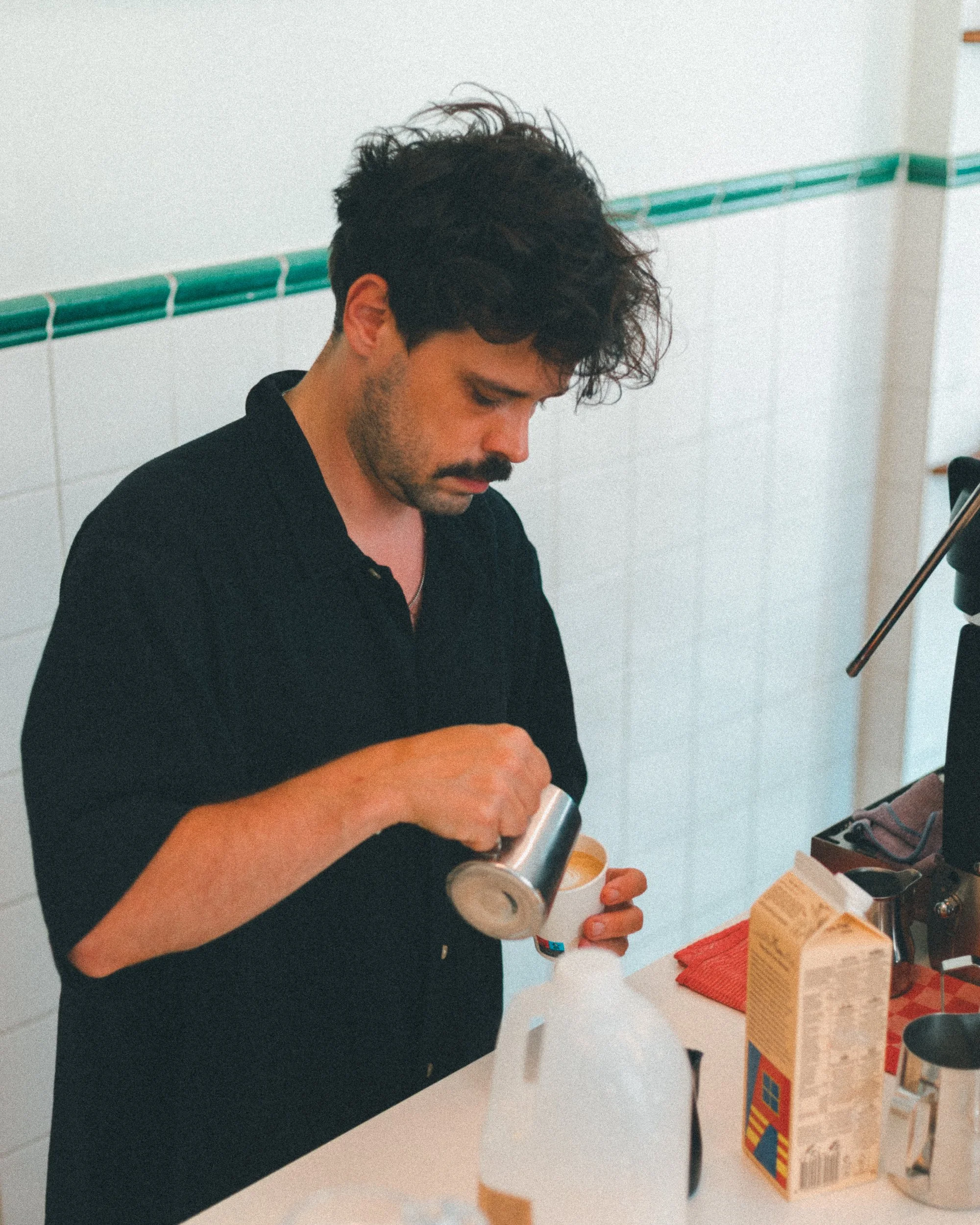 A man with dark curly hair and a mustache is pouring a liquid from a bottle into a small cup at a table with food boxes and kitchen items, in a white-tiled room.
