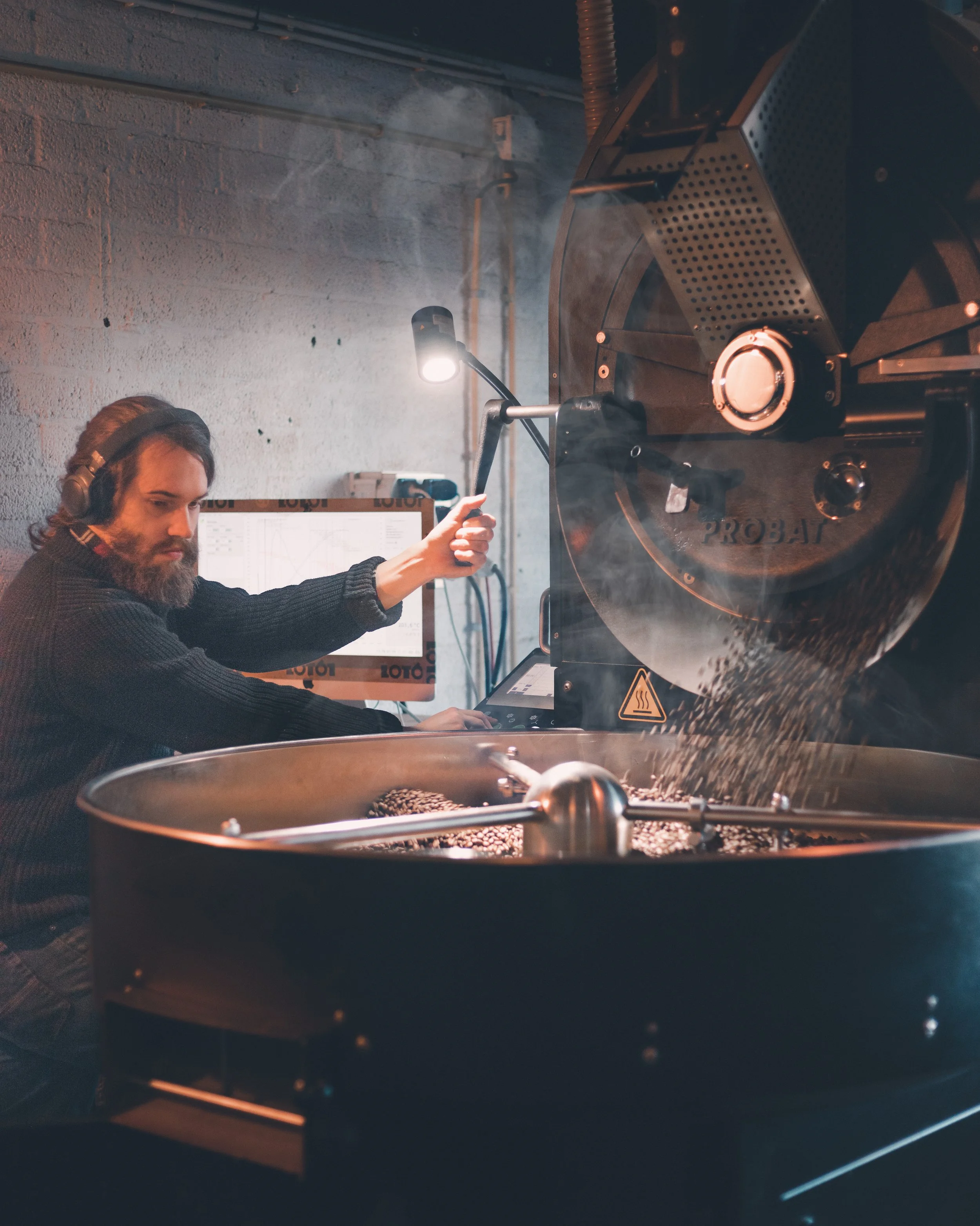 A man with long hair and a beard operating industrial coffee roasting equipment, with a large drum roaster and control panel visible in a dimly lit space.