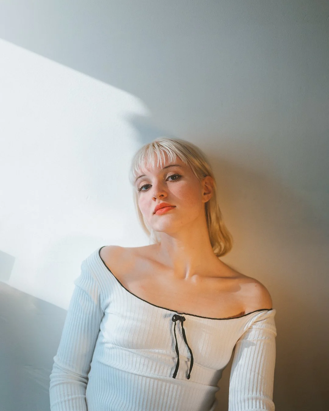 A woman with blonde hair and blue eyes wearing a ribbed, off-the-shoulder top with black trim and a small black bow, standing against a light-colored wall with a soft shadow.