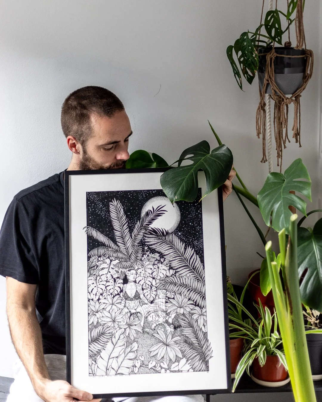 A man holding a framed black and white illustration of a woman surrounded by tropical plants under a starry sky and full moon. There are various houseplants in the background.