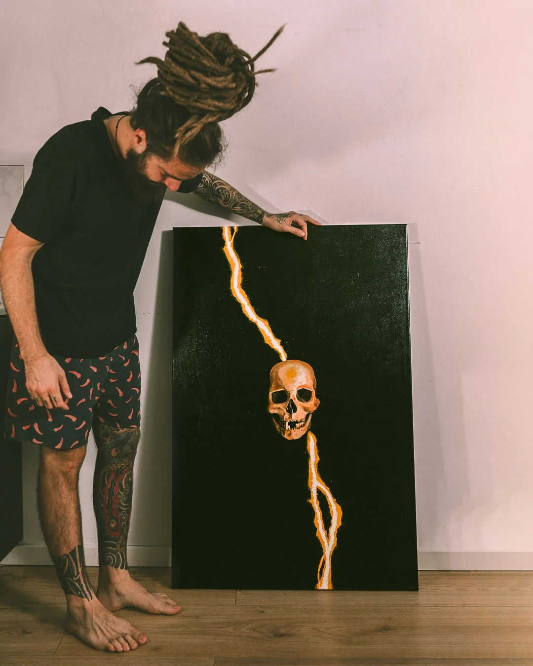 A man with tattoos and dreadlocks looking down at a painting of a skull with lightning bolts on a black background standing against a plain light-colored wall.