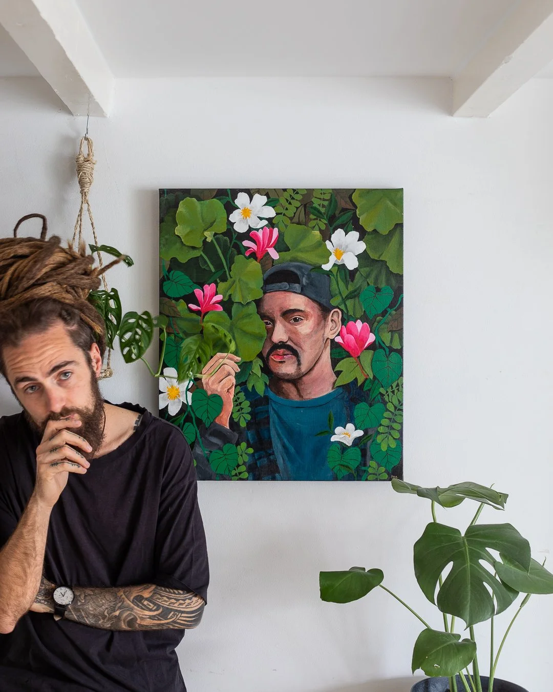 A man with dreadlocks, a beard, and tattoos on his arm, wearing a black shirt, standing in front of a white wall with a colorful portrait of a man with a mustache, wearing a backward cap, surrounded by green leaves and pink and white flowers.
