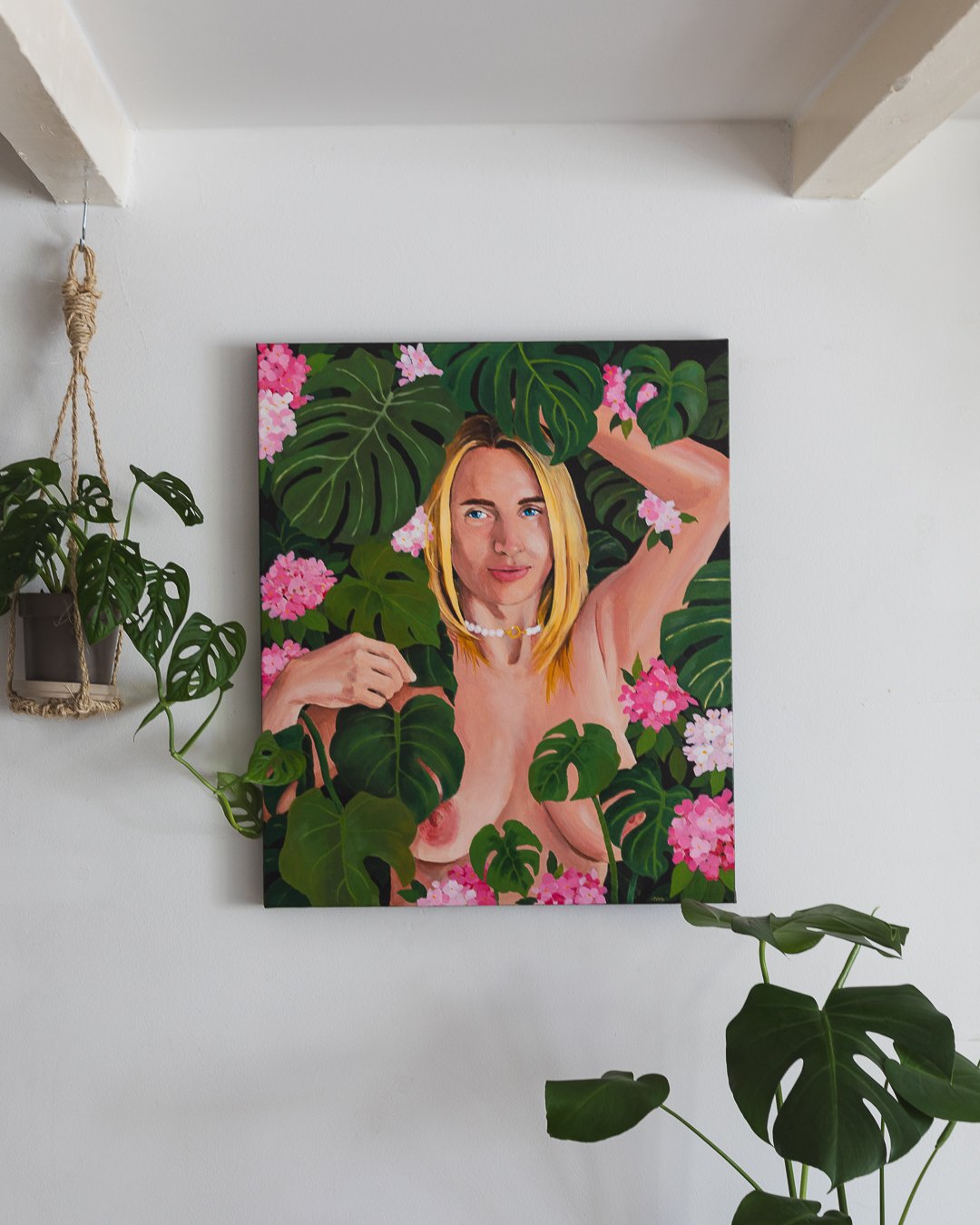 A colorful painting of a blonde woman with blue eyes, surrounded by green leaves and pink flowers, on a white wall.