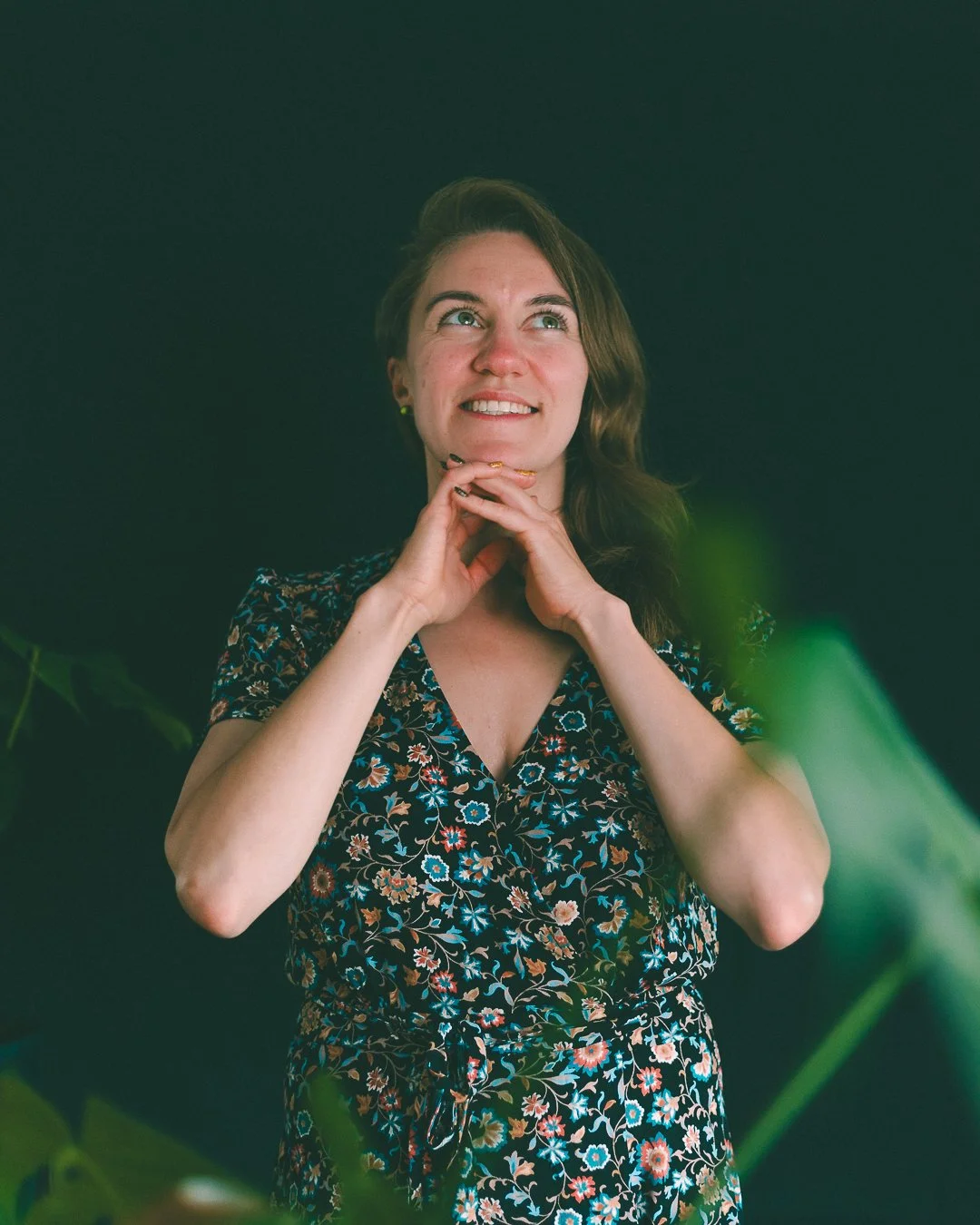 A young woman with long brown hair, wearing a floral dress, is smiling and looking upwards, with her hands clasped under her chin, standing against a dark background surrounded by green leaves.
