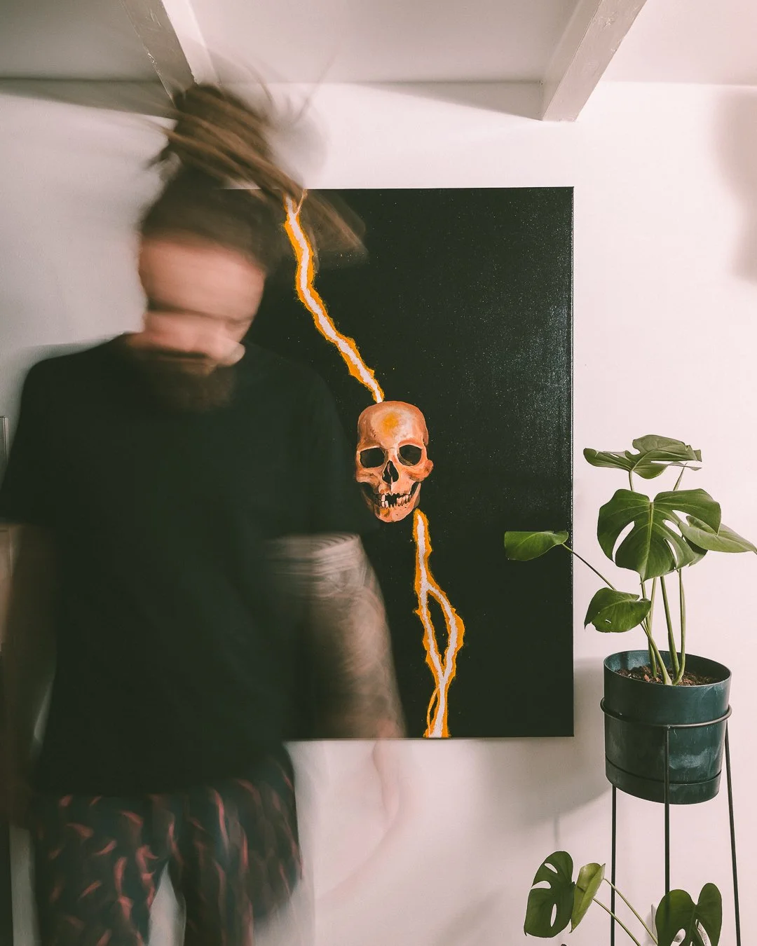 A person with long hair and a beard is moving, causing a blurred effect, standing in front of a painting of a skull with lightning bolts against a black background. There is a potted plant with large leaves on a stand to the right.
