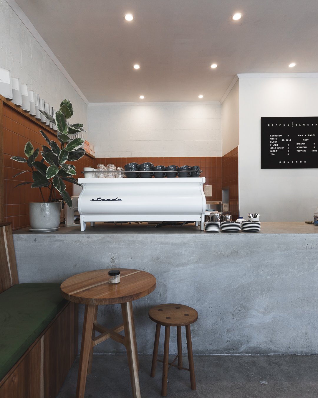 Interior of a coffee shop with a white espresso machine on a concrete counter, a tall potted plant, a small wooden table with a small jar, a wooden stool, and a black menu board on the wall.