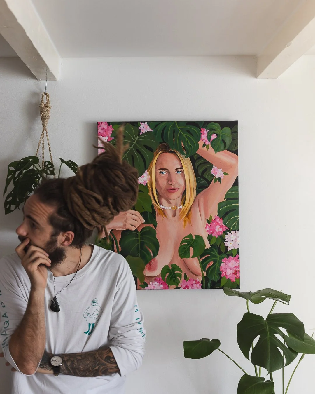 A man with dreadlocks and tattoos on his arm is standing in front of a colorful portrait of a woman with blonde hair, green eyes, and a pearl necklace, surrounded by green leaves and pink flowers.