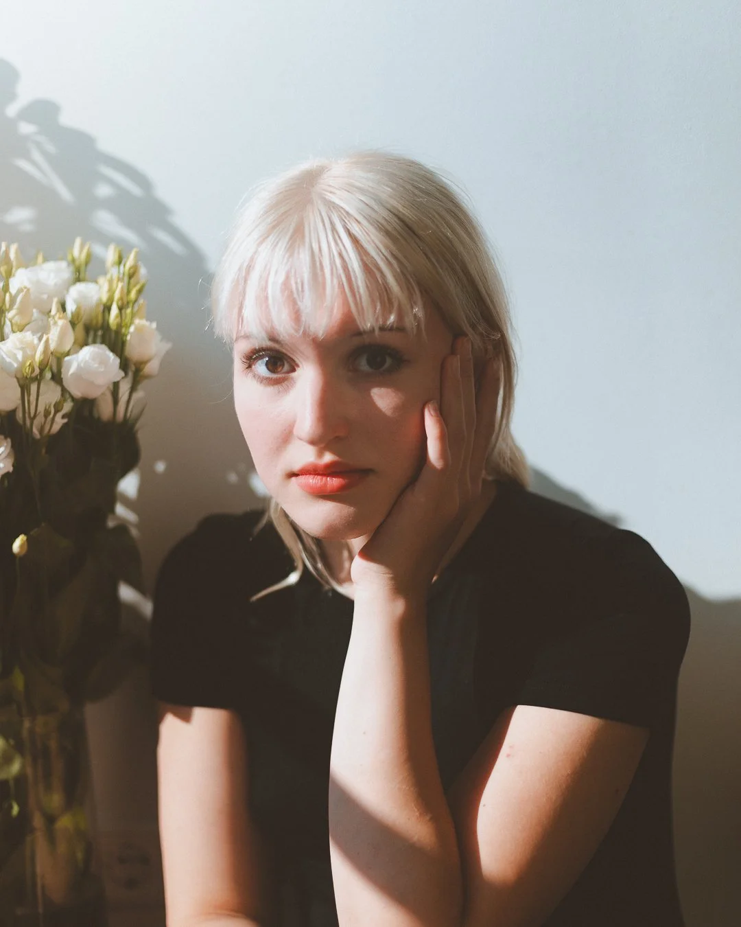 A young woman with blonde hair and light skin resting her face on her hand, sitting next to a bouquet of white flowers against a plain background.