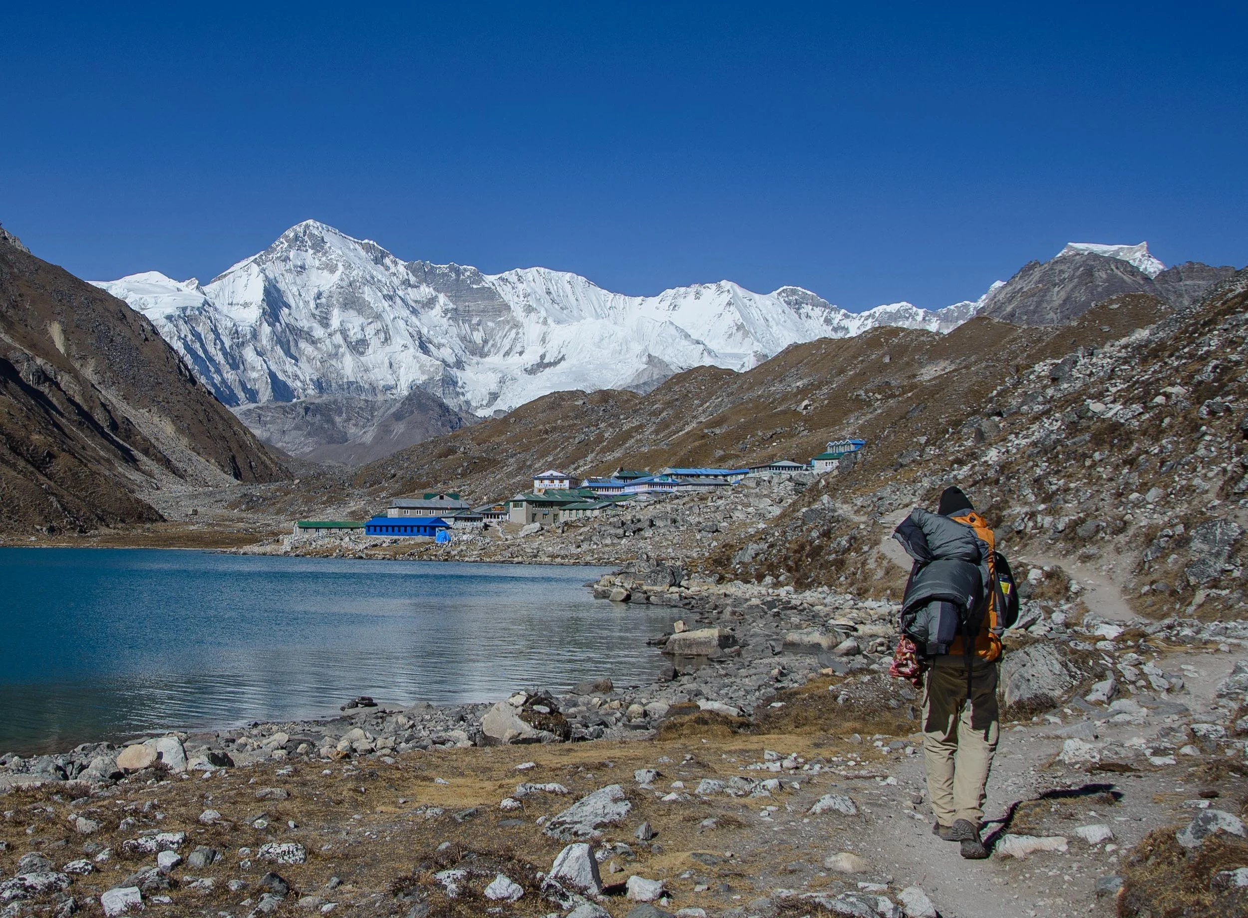 A trekker walking beside Gokyo Lake toward Gokyo village, with the massive Cho Oyu rising in the background.