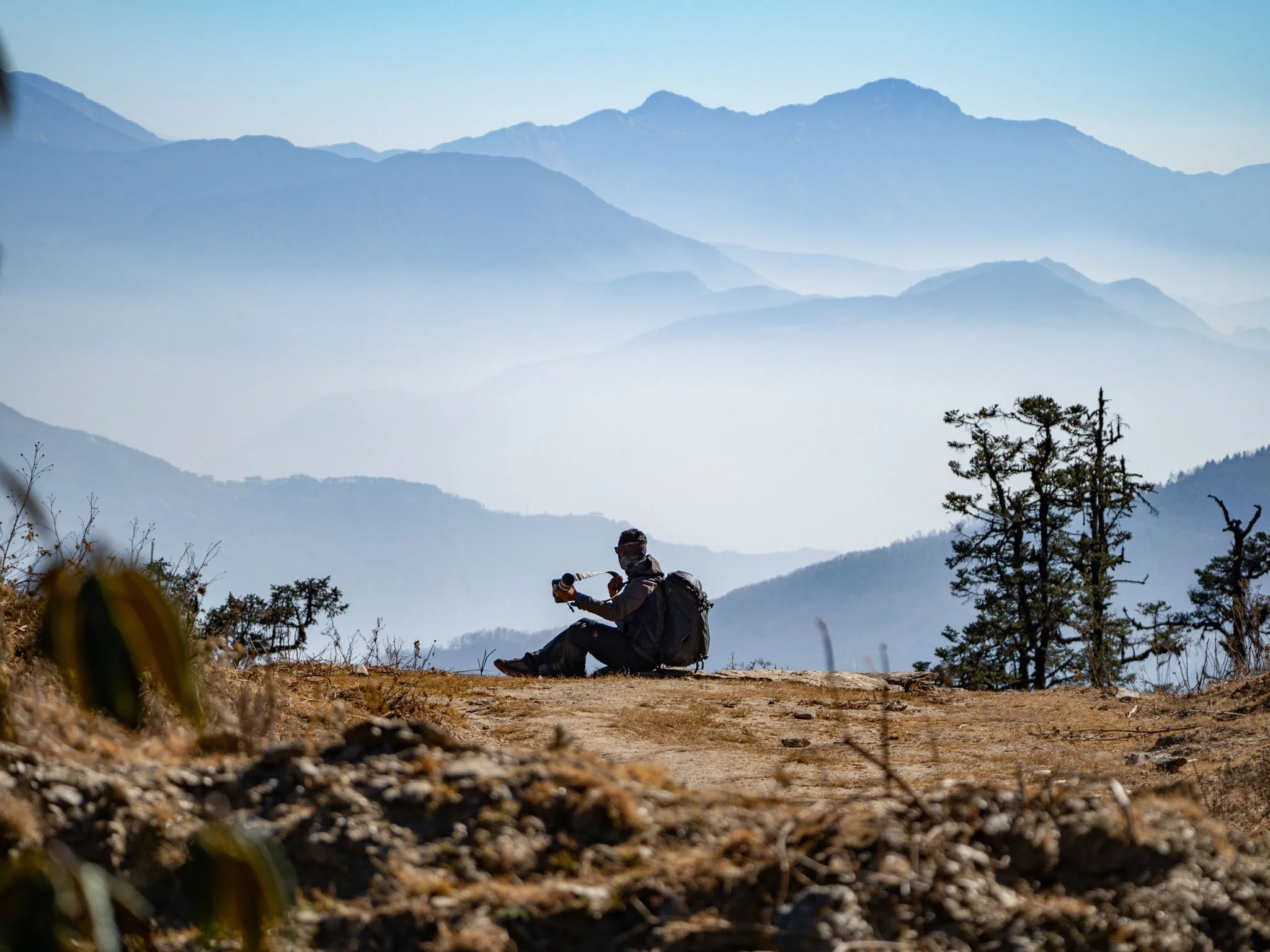 Person taking a photo in a mountainous landscape with misty hills and sparse trees, under a clear blue sky.